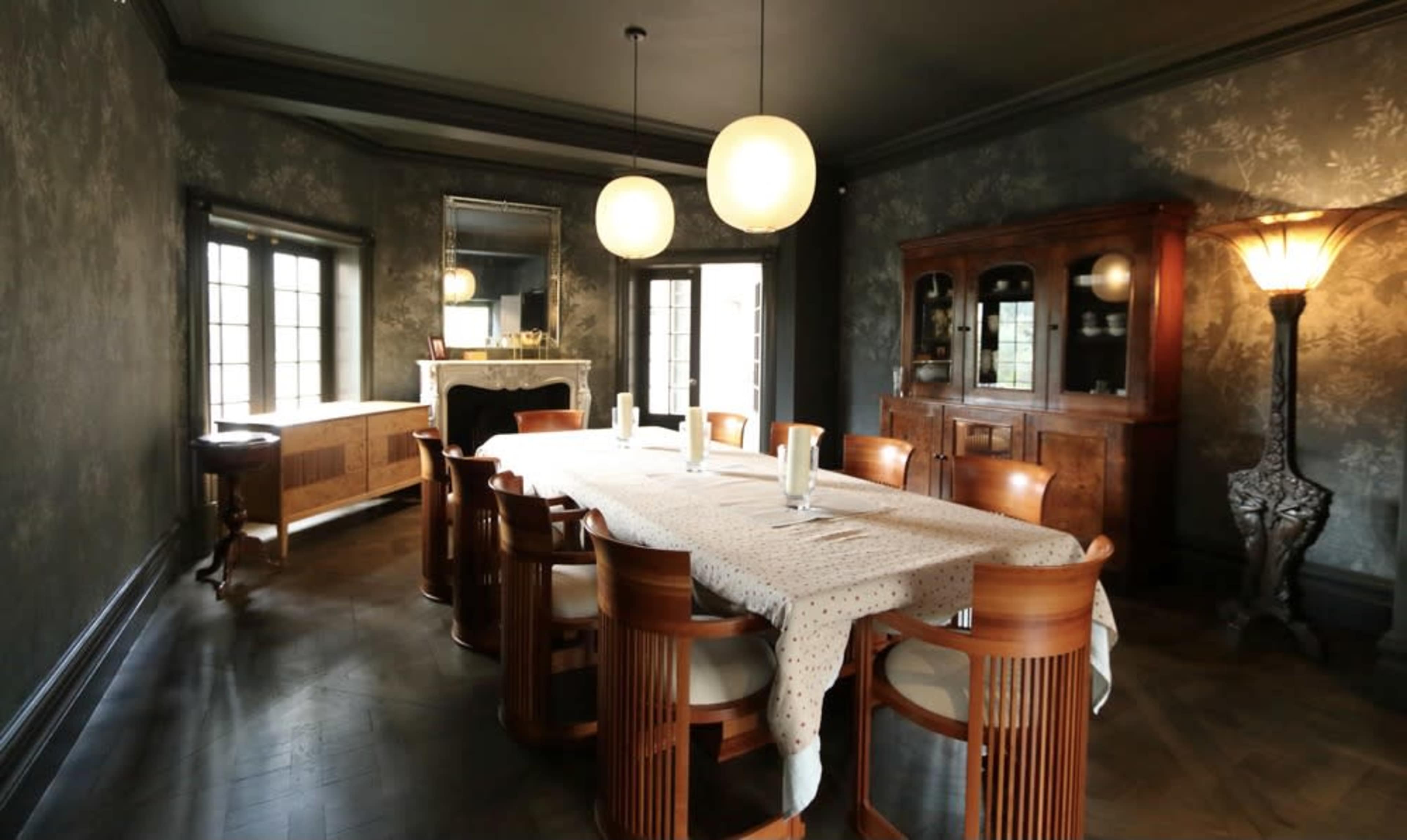 A spacious dining room features a long table surrounded by wooden chairs, with a decorative cabinet and mirror against the walls.