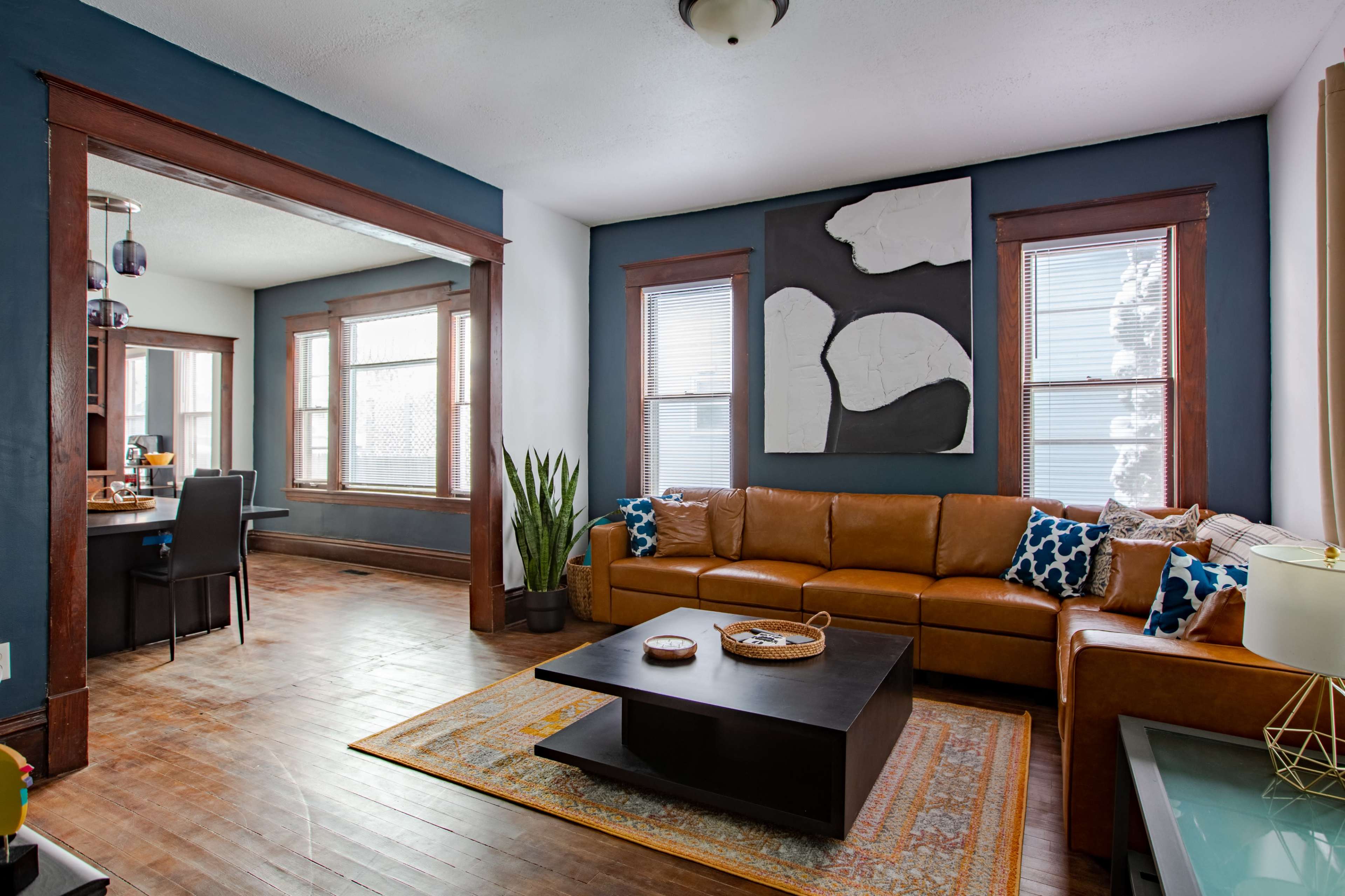 The image shows a living room with a brown leather sectional sofa, a black coffee table, and a large abstract wall decoration, with a dining area visible through an archway.