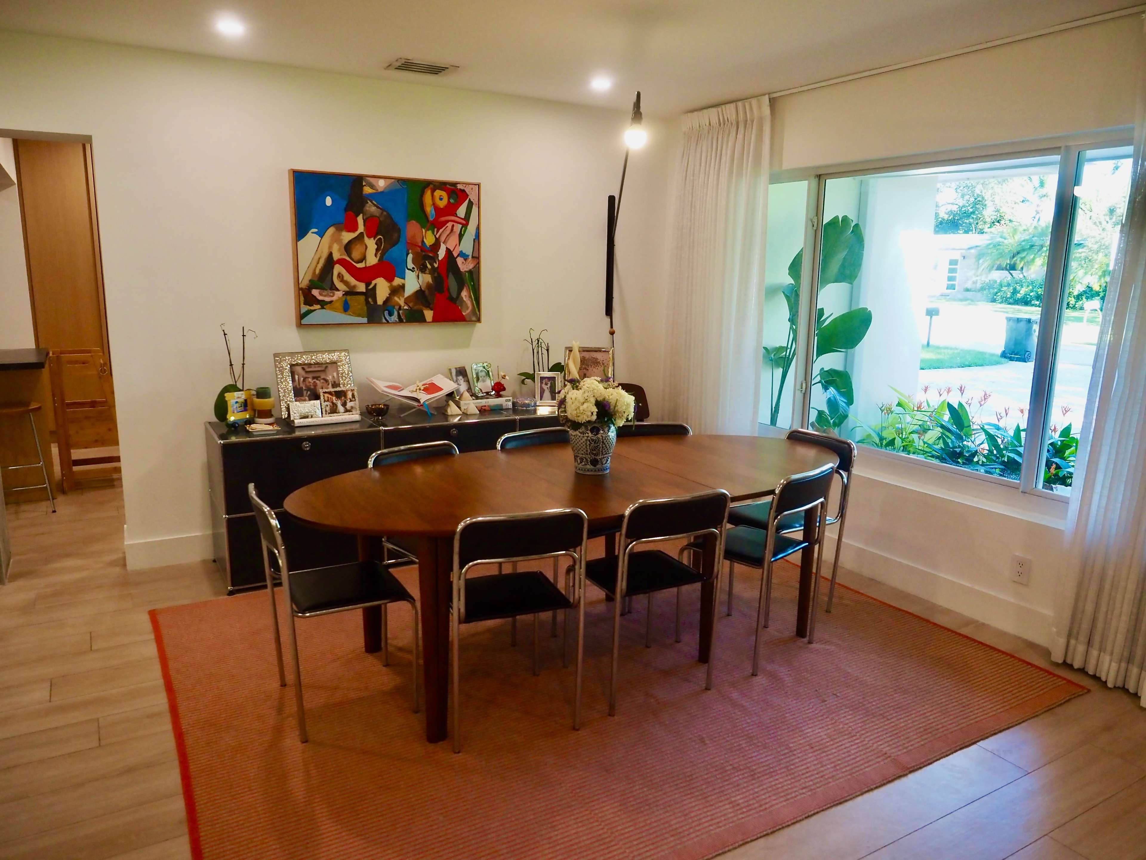 A dining area features a circular wooden table surrounded by six metal chairs, with a large window overlooking greenery and an abstract painting on the wall.