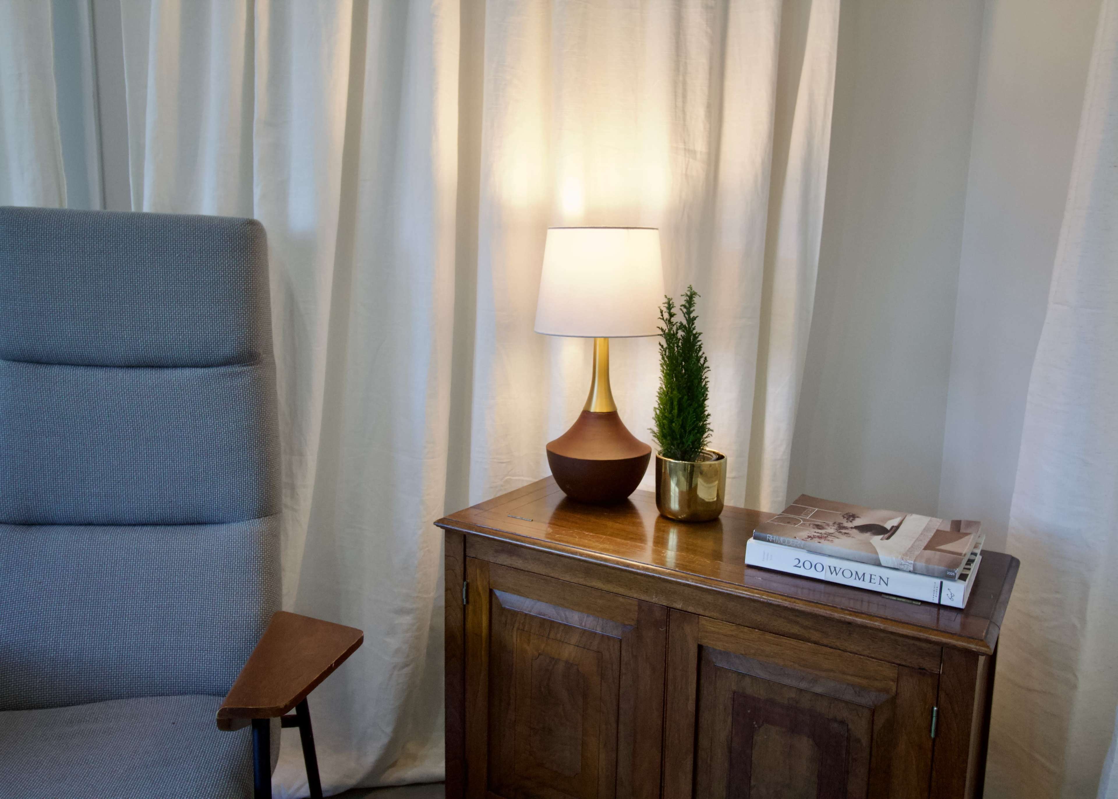 A modern lamp and a potted plant are placed on a wooden side table beside a tall chair, with flowing white curtains in the background.