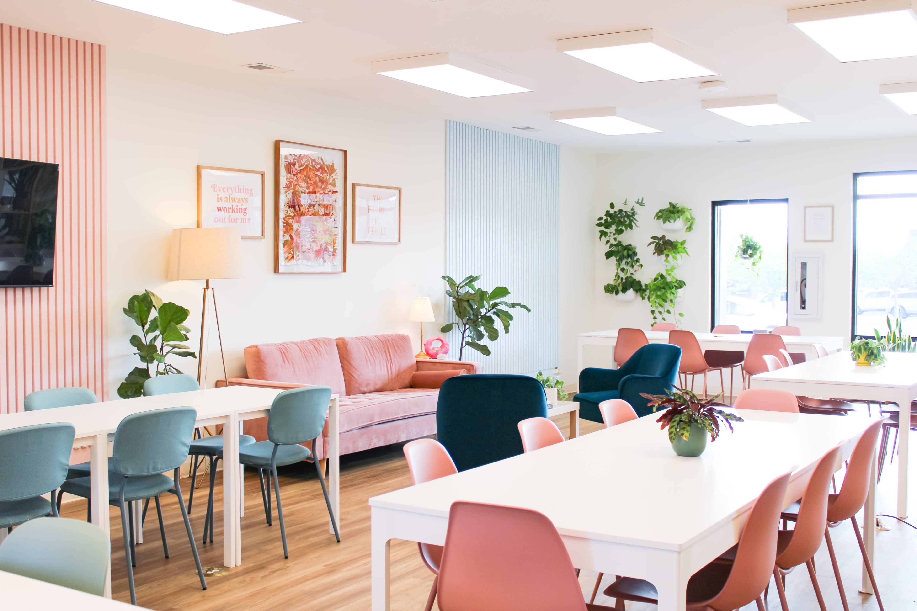 A bright and modern cafe or communal workspace features pastel-colored chairs, white tables, potted plants, and a pink sectional sofa.