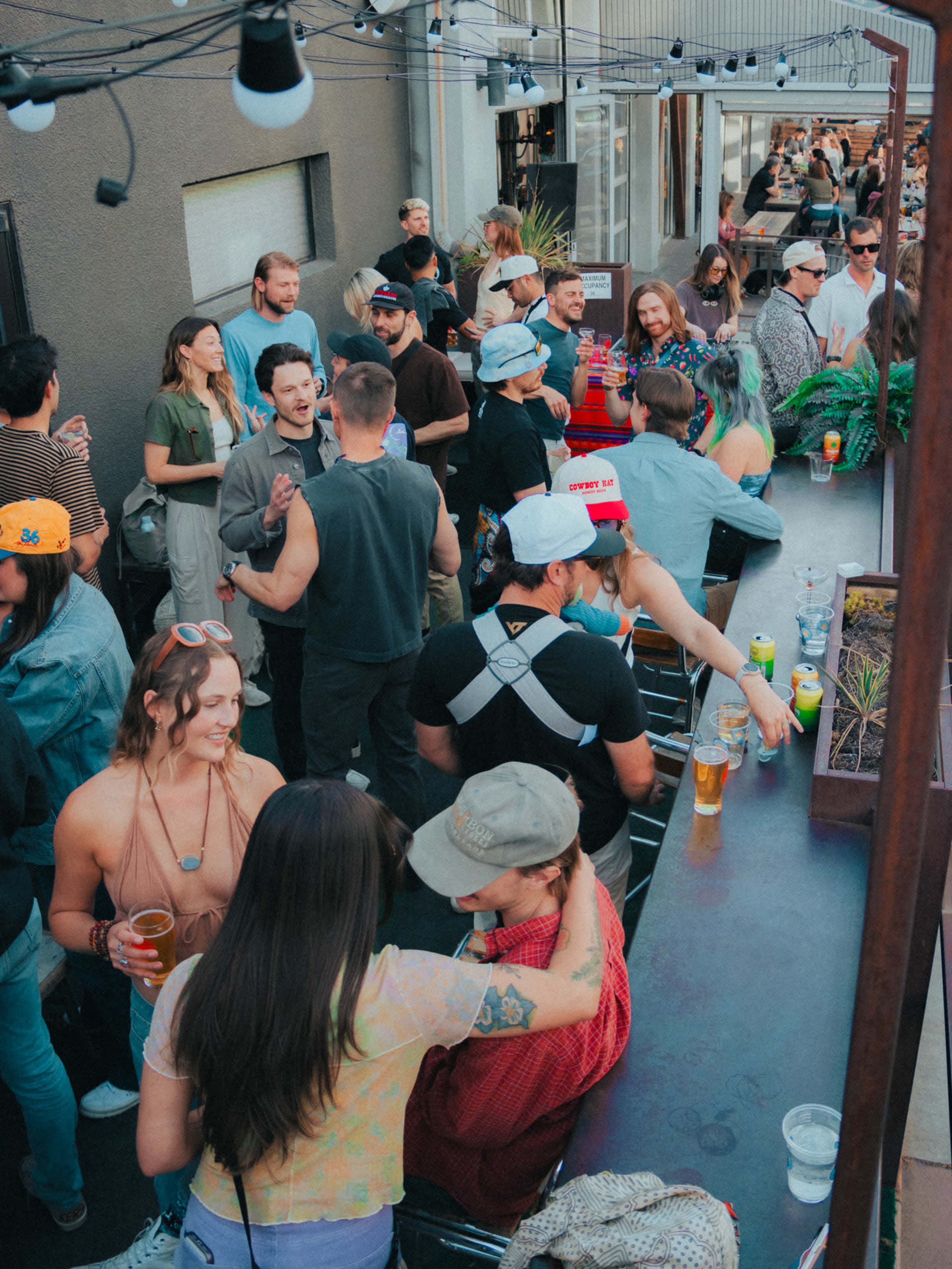 A crowded rooftop bar scene features people socializing, drinking, and enjoying the atmosphere on a warm day.