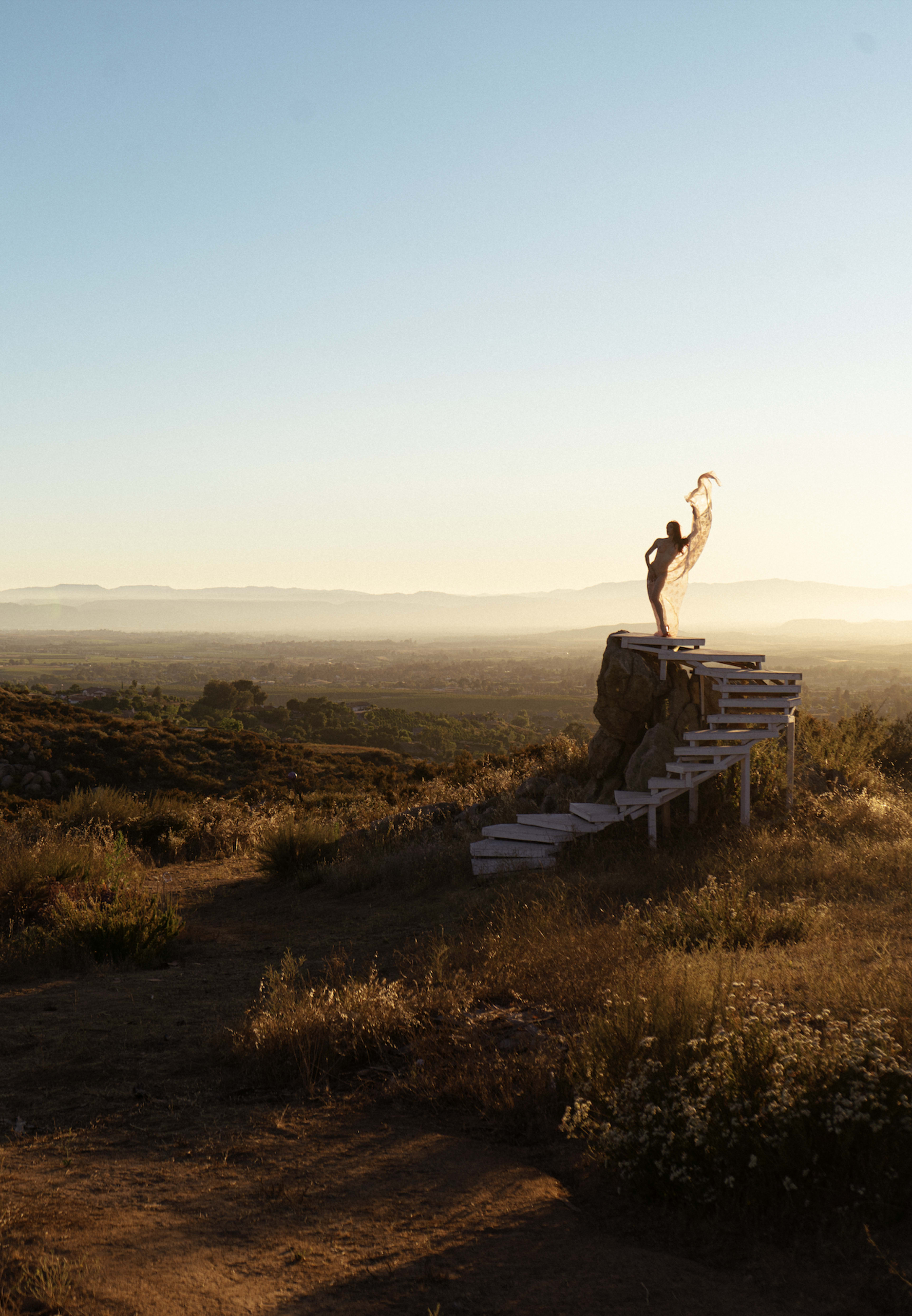 A figure stands on a rocky outcrop, draped in a flowing fabric, overlooking a vast landscape during sunset.