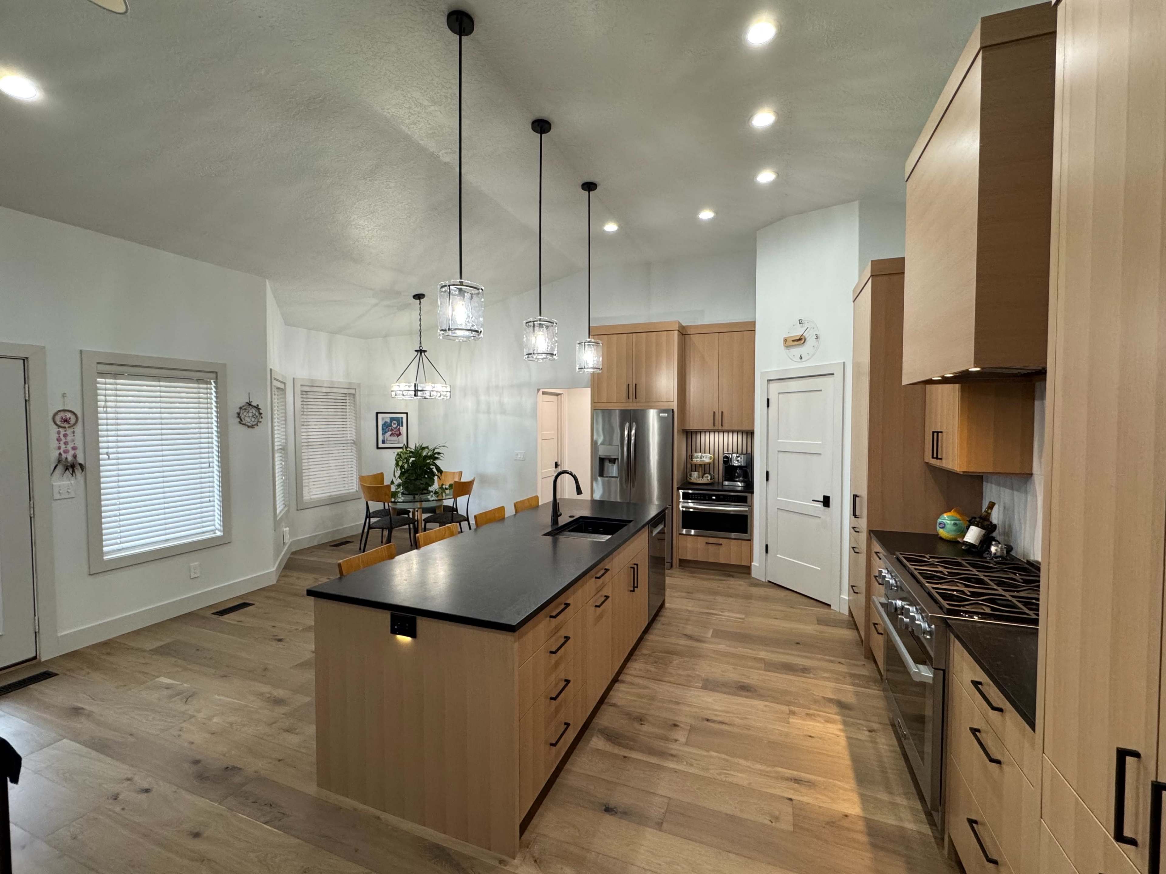 The image shows a modern kitchen with wood cabinetry, a central island with a black countertop, and a dining area in the background.