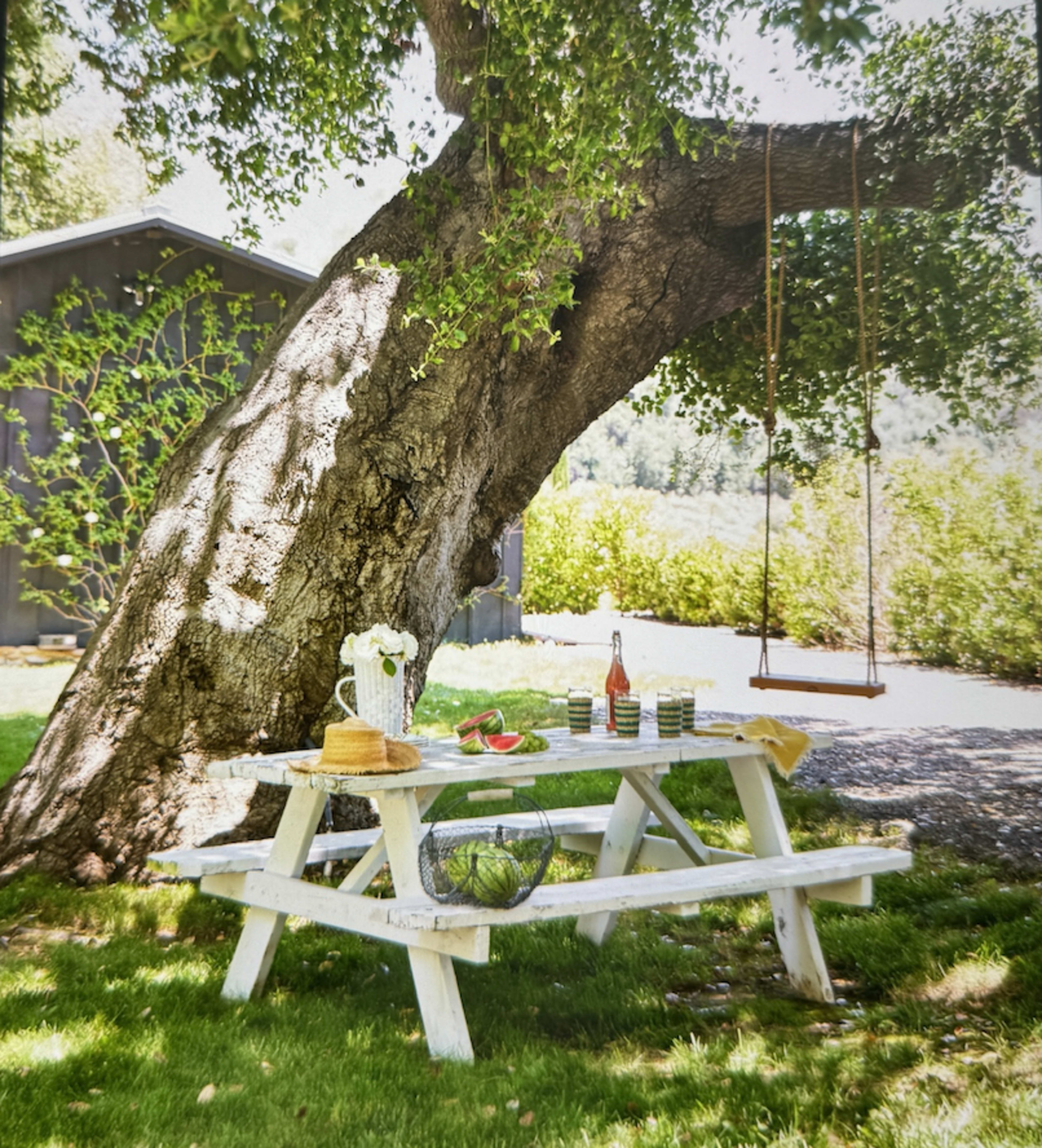 A white picnic table with a checkered cloth is set under a large tree, accompanied by bottles and bowls of food, while a swing hangs from the tree's branch.