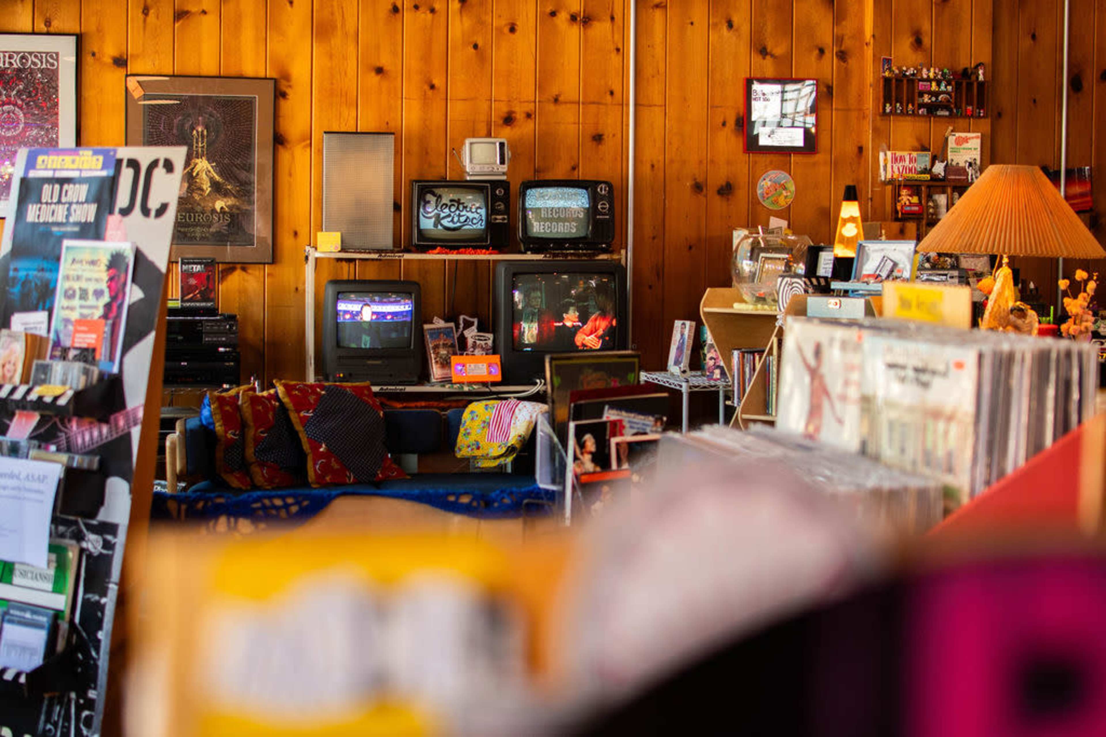 The image shows a cozy vintage store interior with wooden walls, featuring multiple old televisions displaying a performance and stacks of vinyl records.