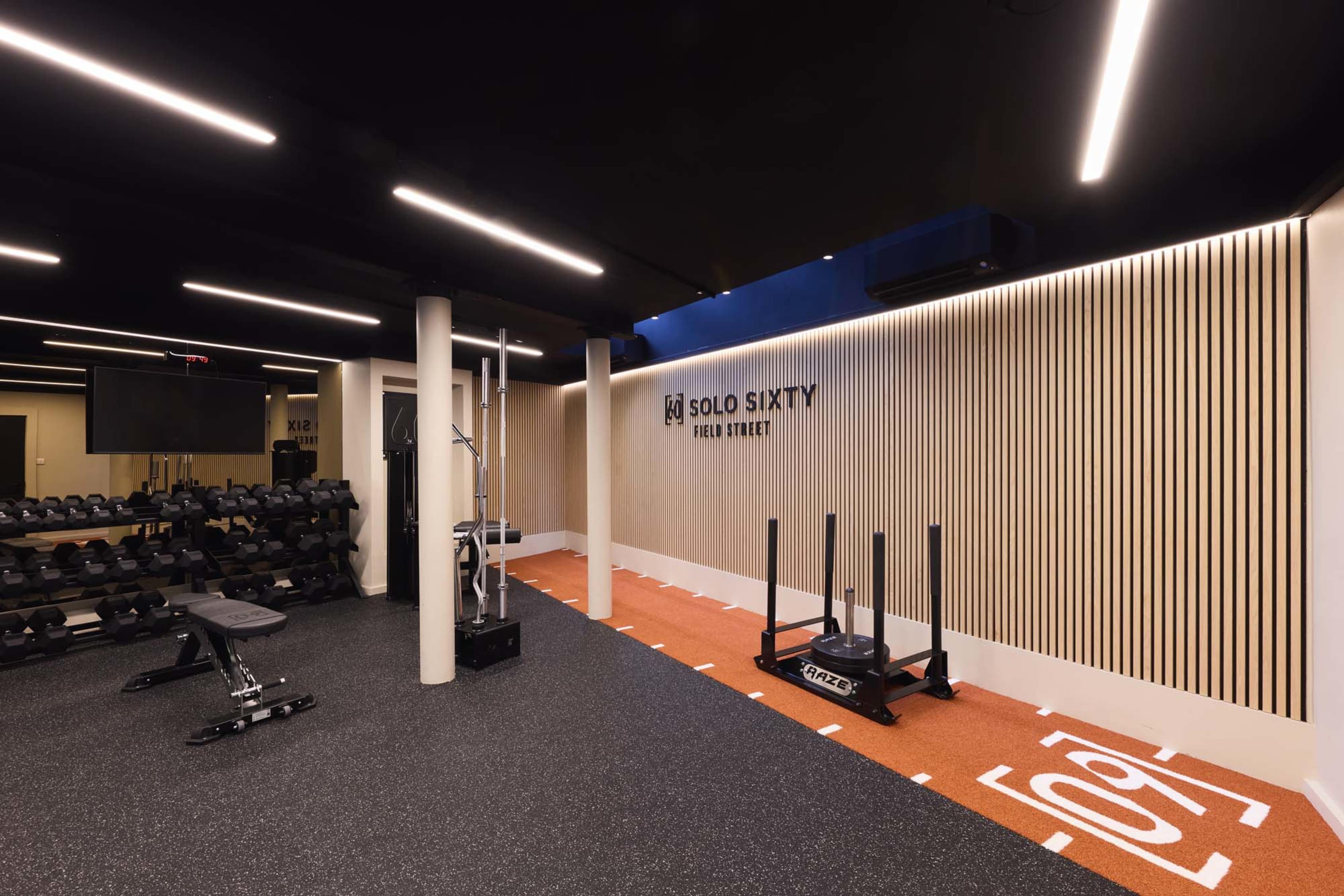 The image shows a modern gym interior with weightlifting equipment, a sled, and a designated exercise area featuring a turf surface.