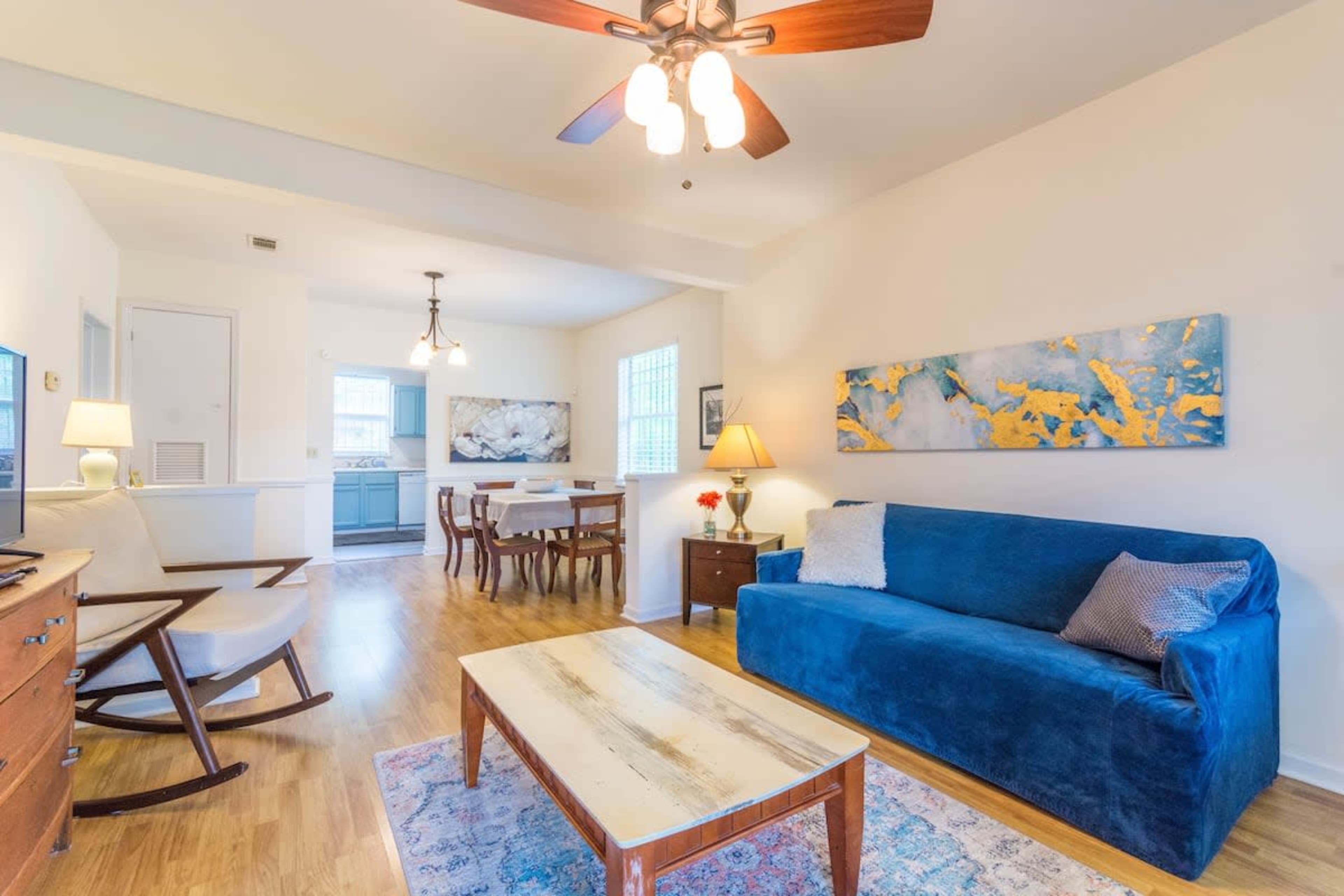 A bright living room with a blue sofa, a wooden coffee table, and a dining area in the background.