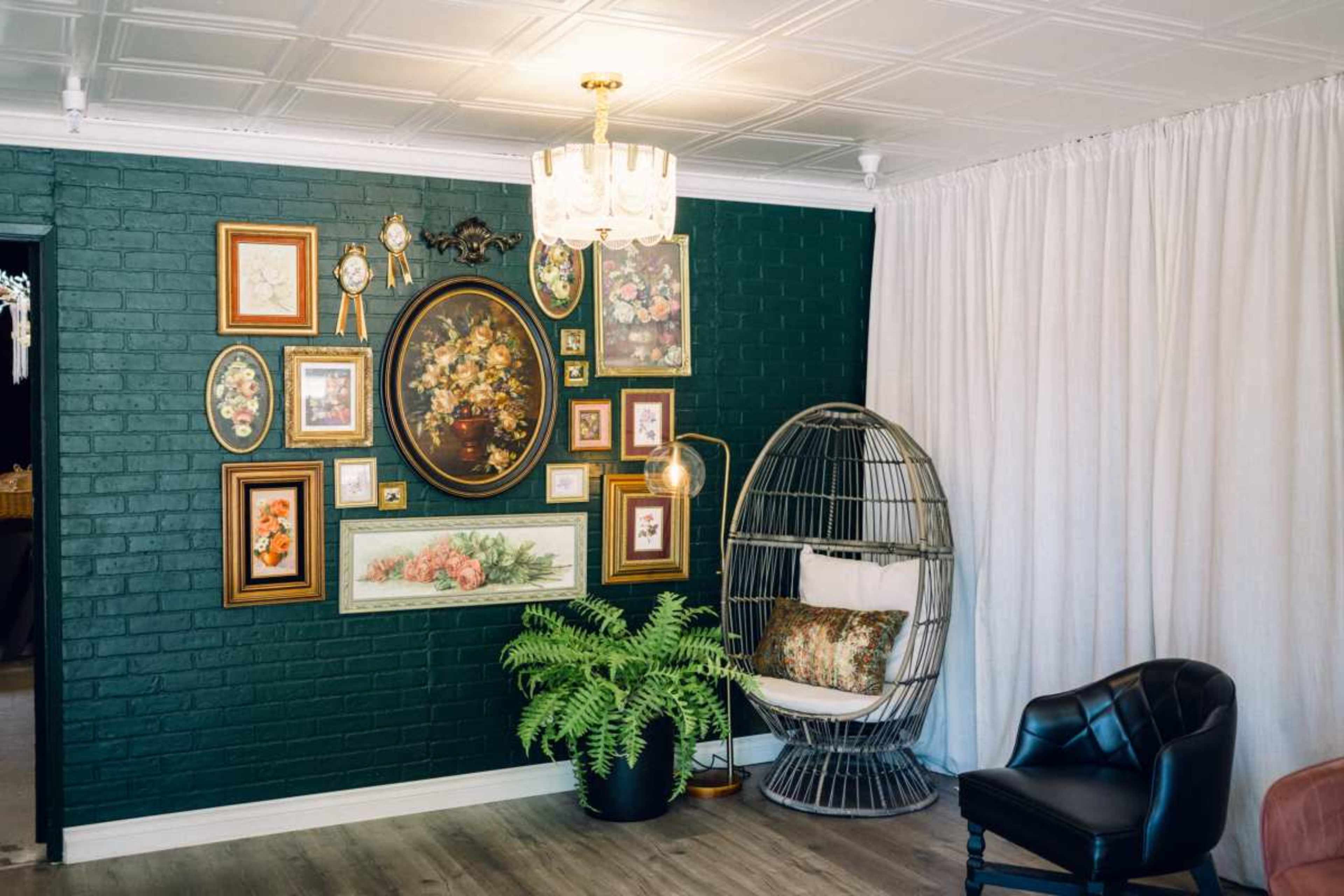 The image shows a living room with a dark green brick wall adorned with various framed artworks, a hanging chair, and a potted fern.
