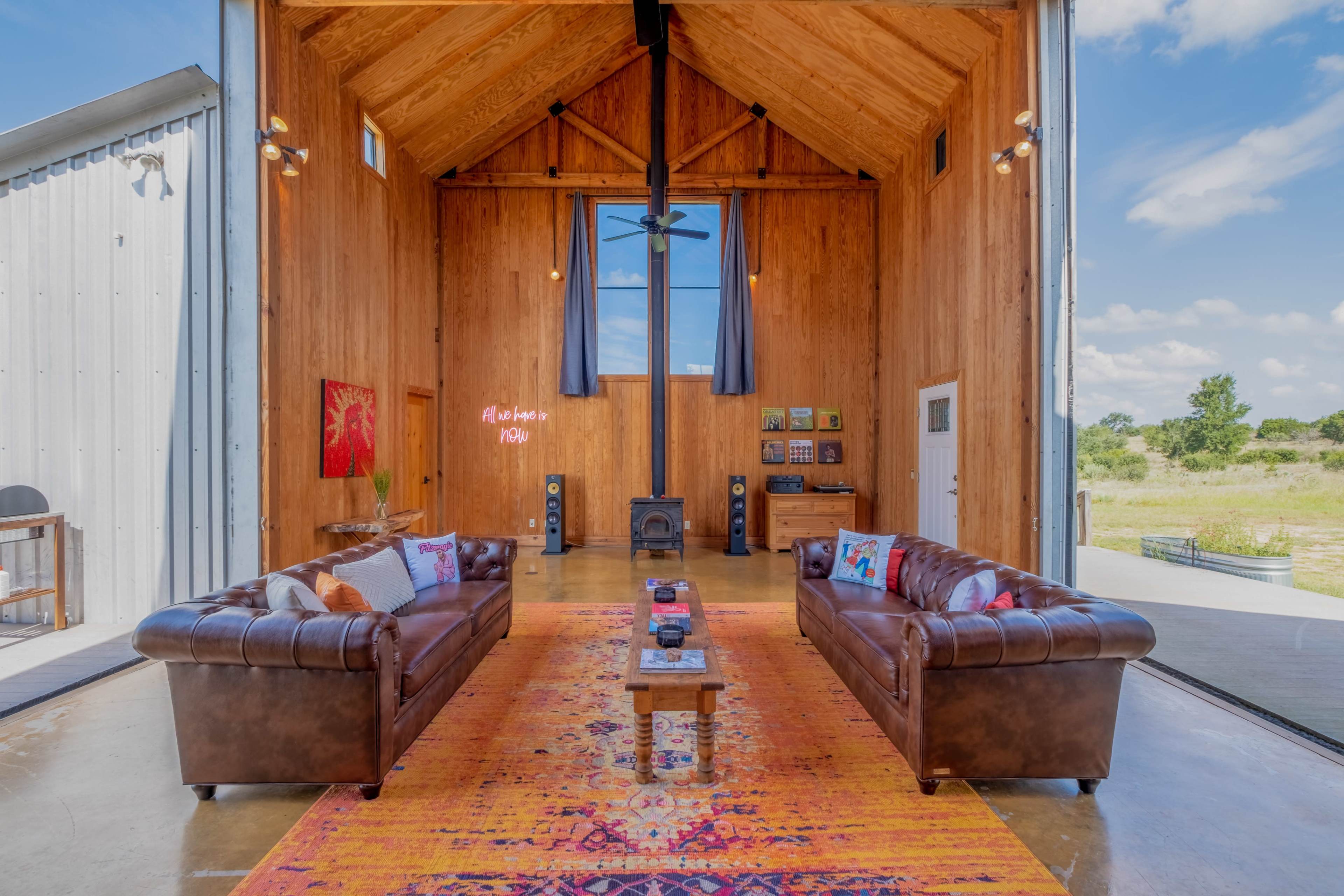 A spacious, rustic interior features two leather sofas facing a central coffee table on a vibrant rug, with large windows allowing natural light to fill the room.