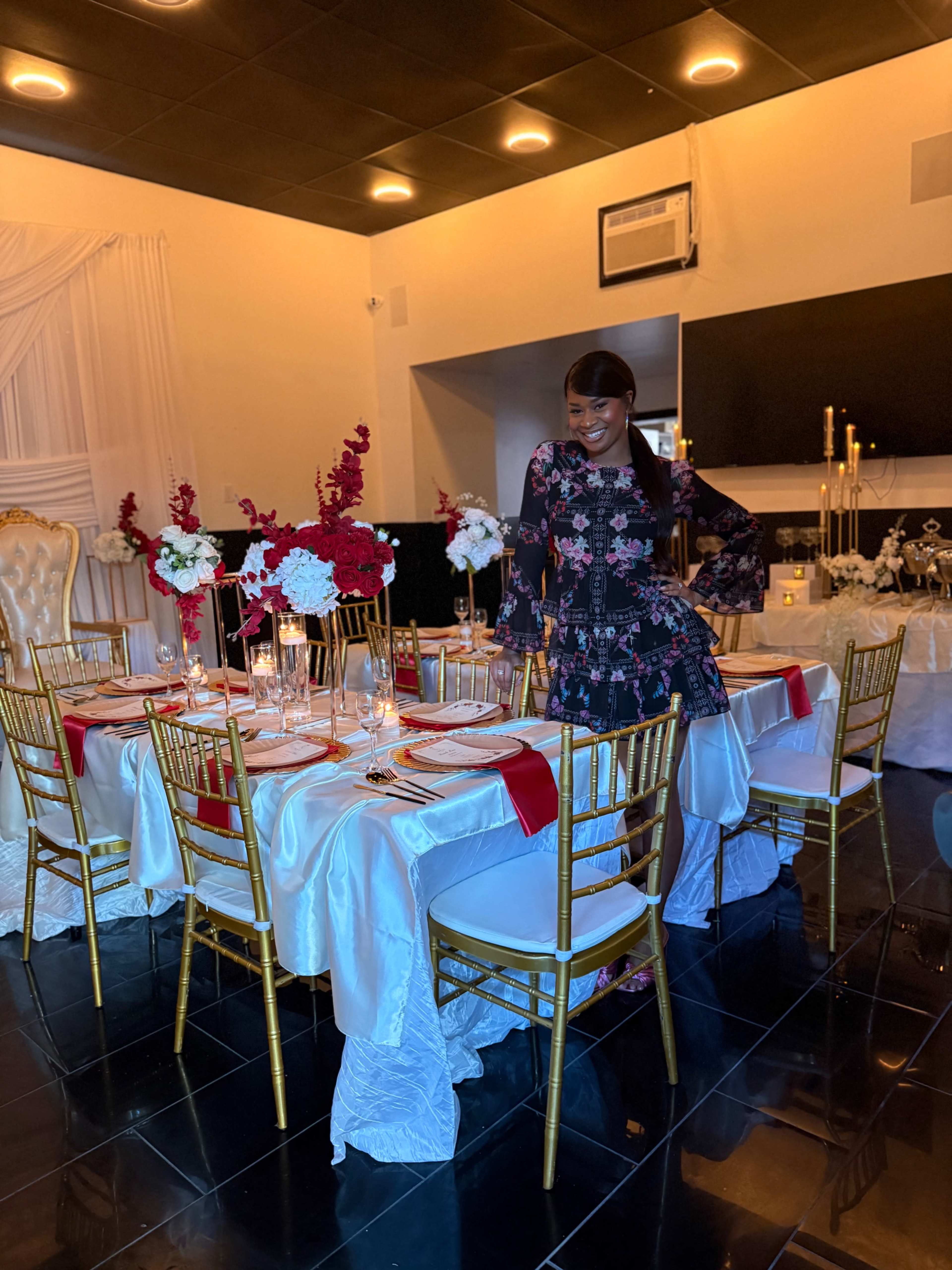 A woman stands next to an elegantly set dining table adorned with floral centerpieces and gold chairs in a well-decorated room.