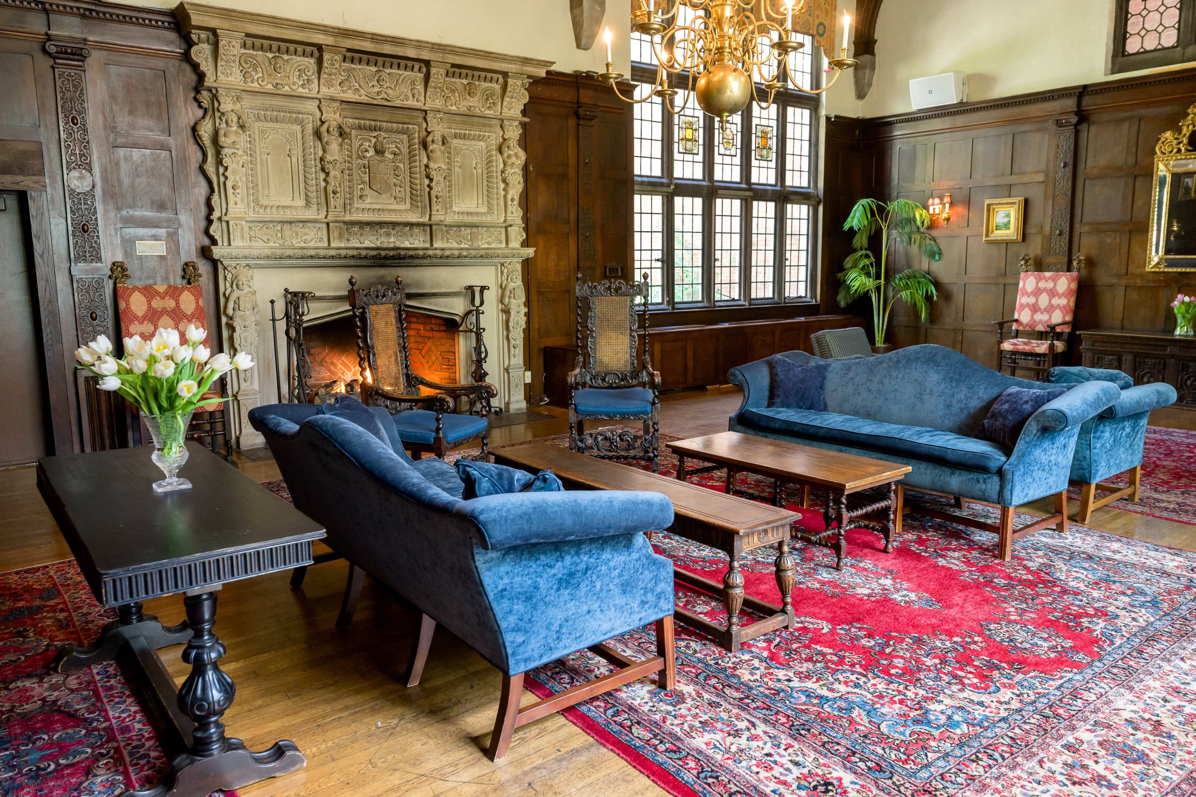 A spacious living room featuring blue upholstered sofas, a wooden coffee table, a large fireplace, and intricate wall paneling, all set on a decorative area rug.