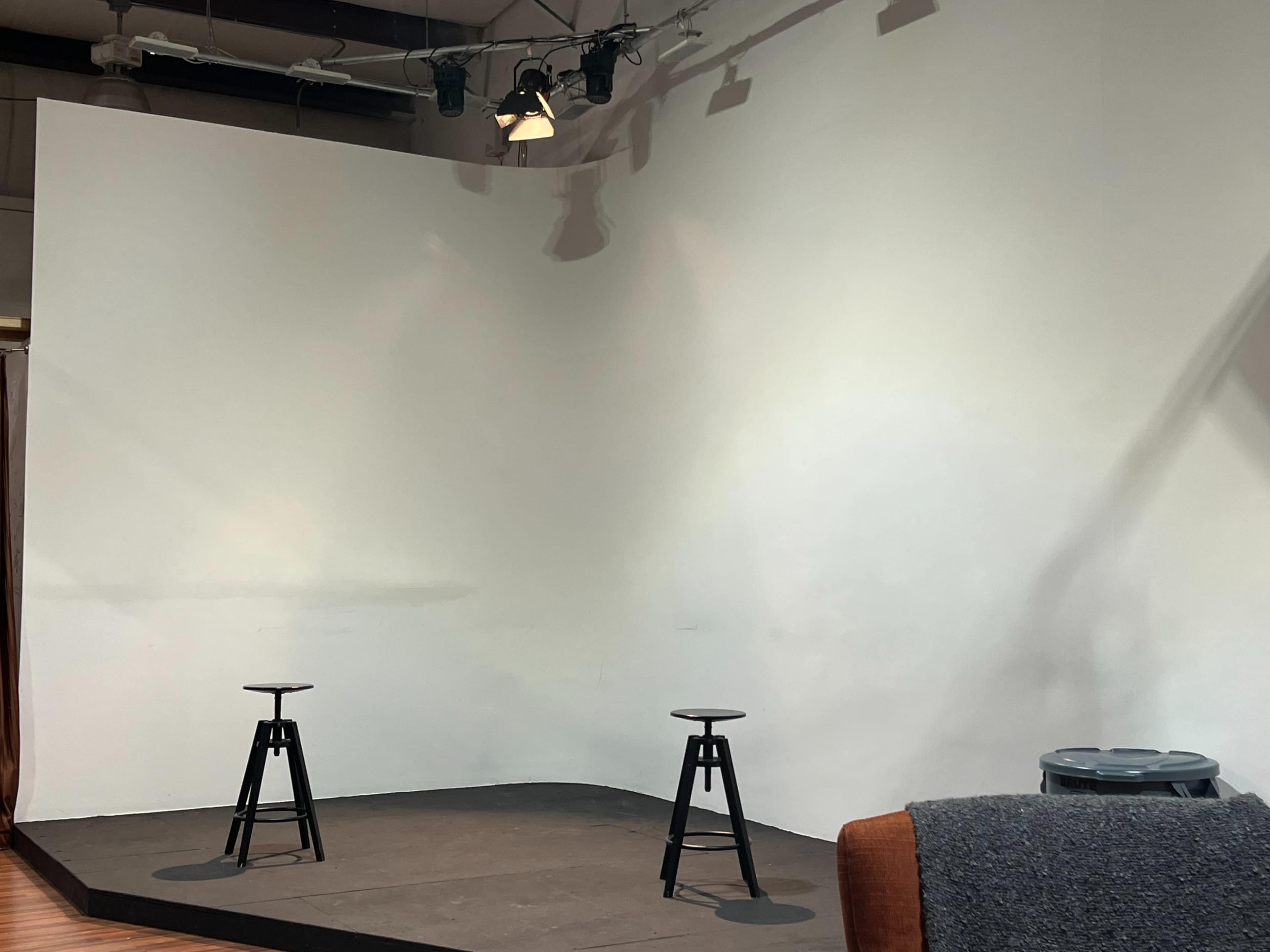 A minimalist stage setup featuring two black stools on a wooden floor in front of a blank white wall.
