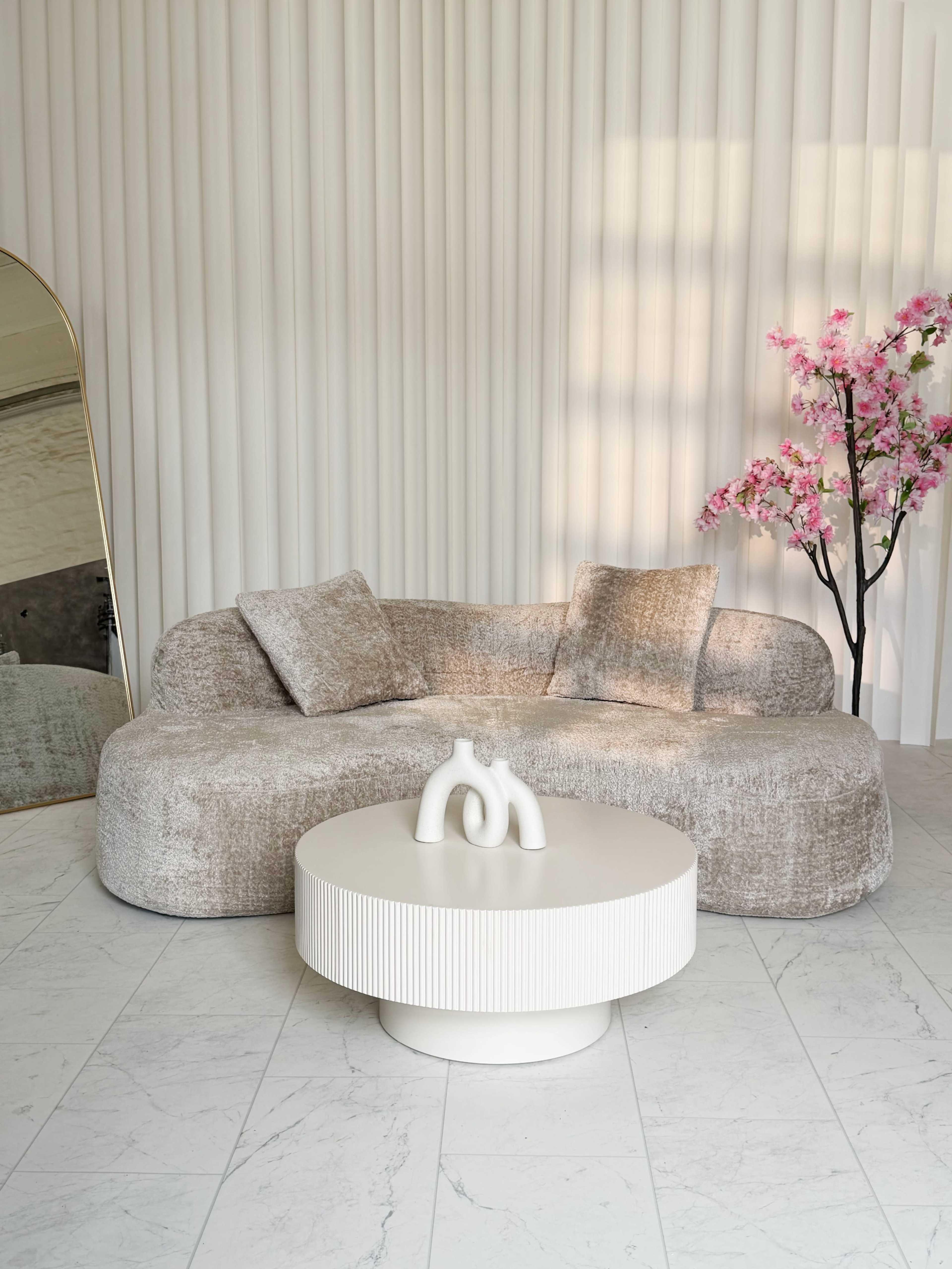A curved, beige sofa is positioned next to a round, white coffee table, with a decorative vase on top, in a room featuring vertical white paneling and a faux cherry blossom tree.