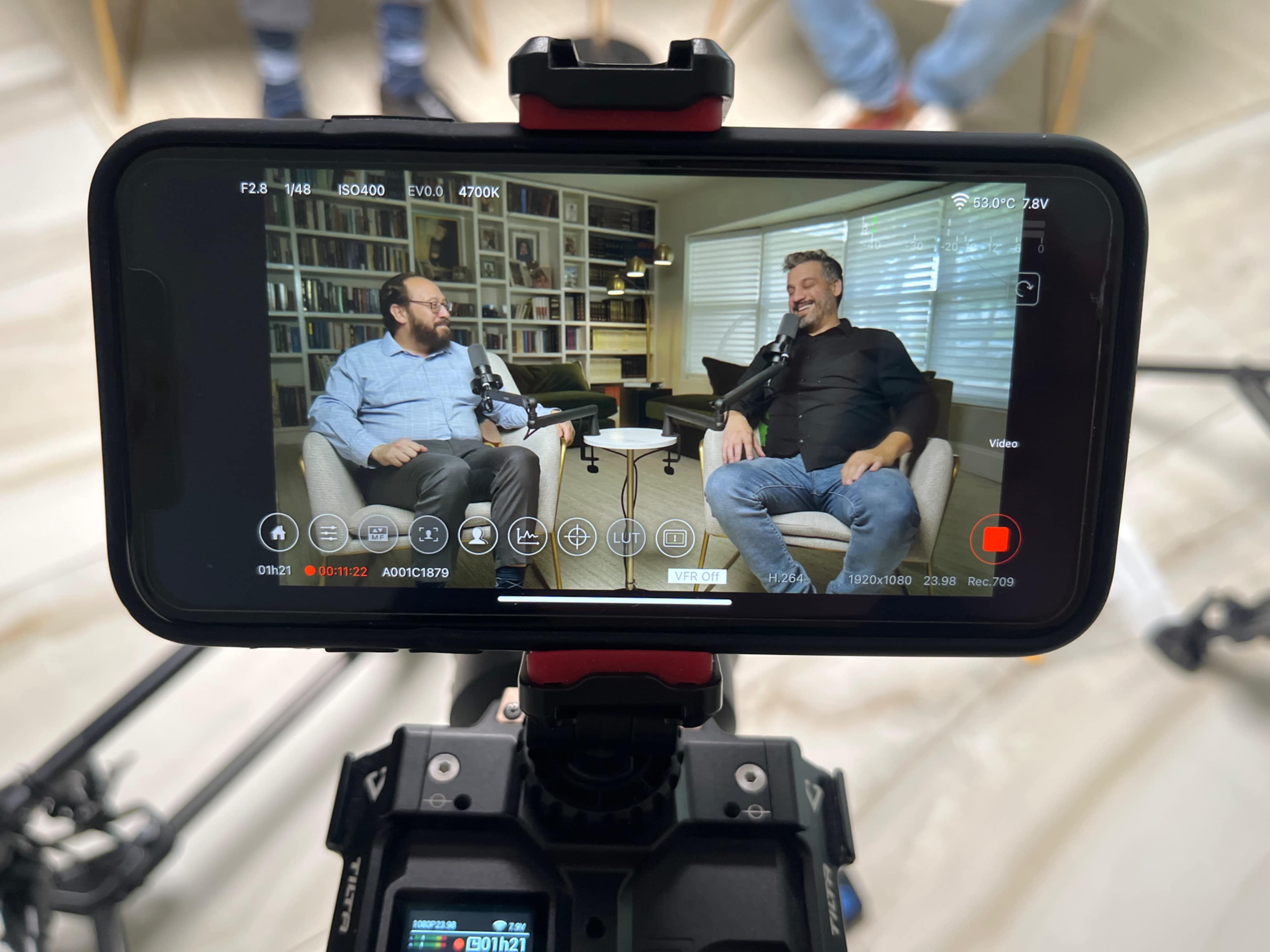 A smartphone camera captures two men seated and conversing in an interview setting, with microphones positioned for recording.