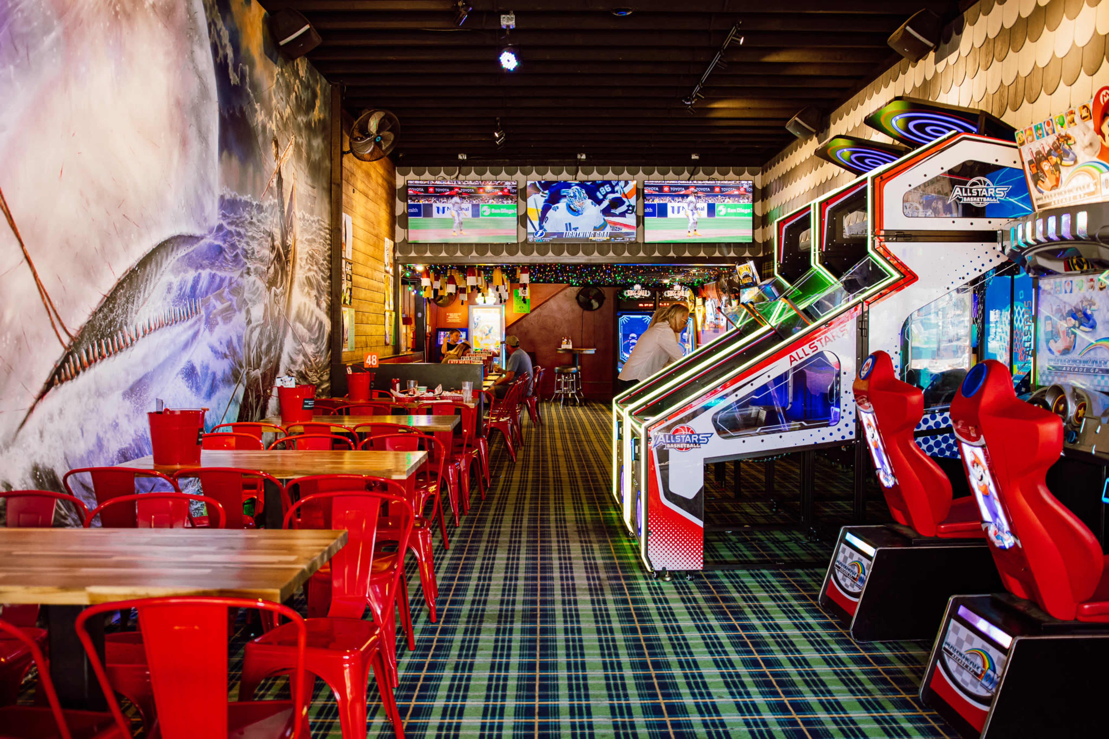 The image shows a colorful arcade featuring several gaming machines, tables with red chairs, and a wall mural depicting a wave.