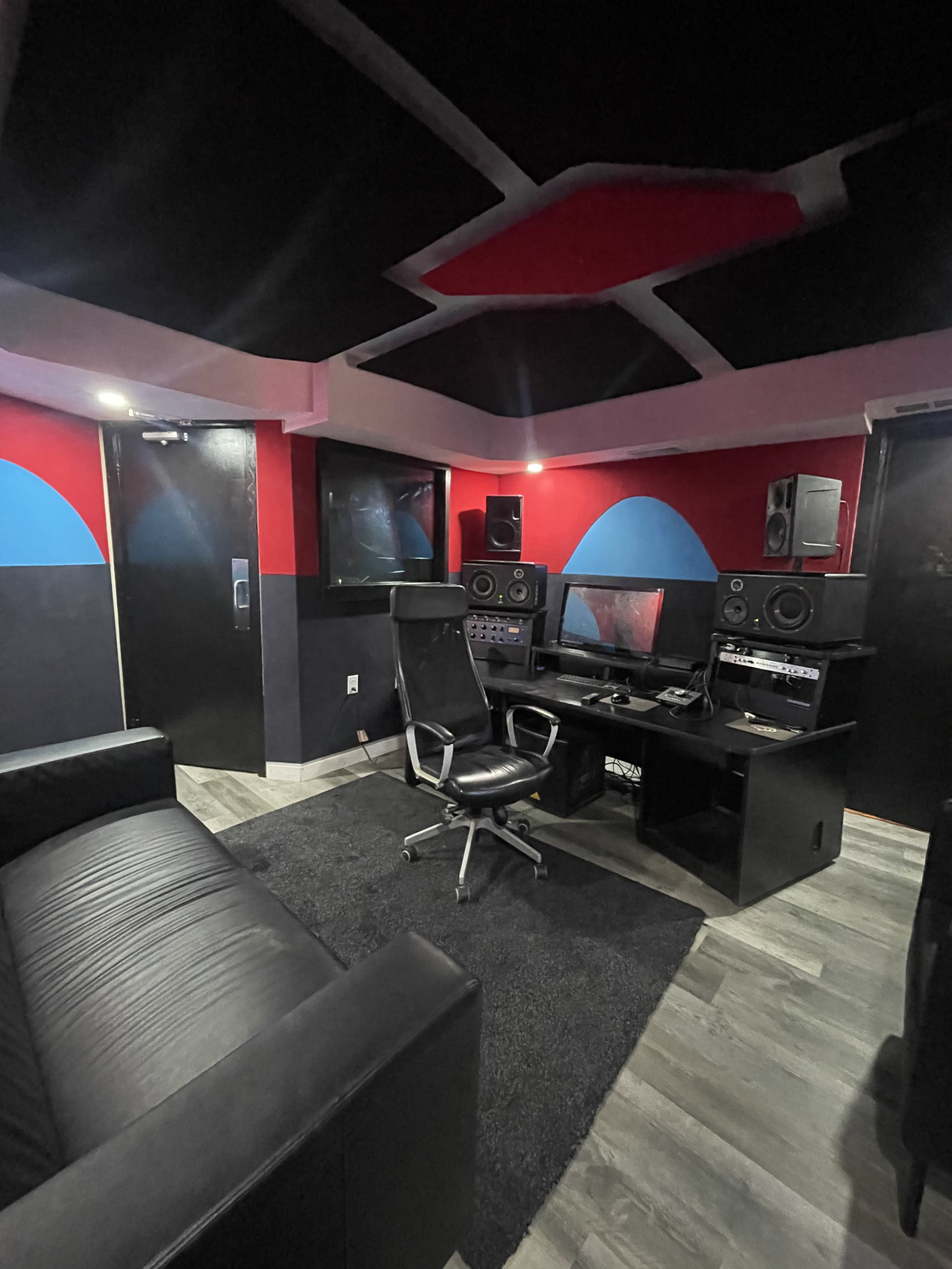 The image shows a dimly lit recording studio featuring a black leather couch, a desk with audio equipment, and soundproofing materials on the walls.