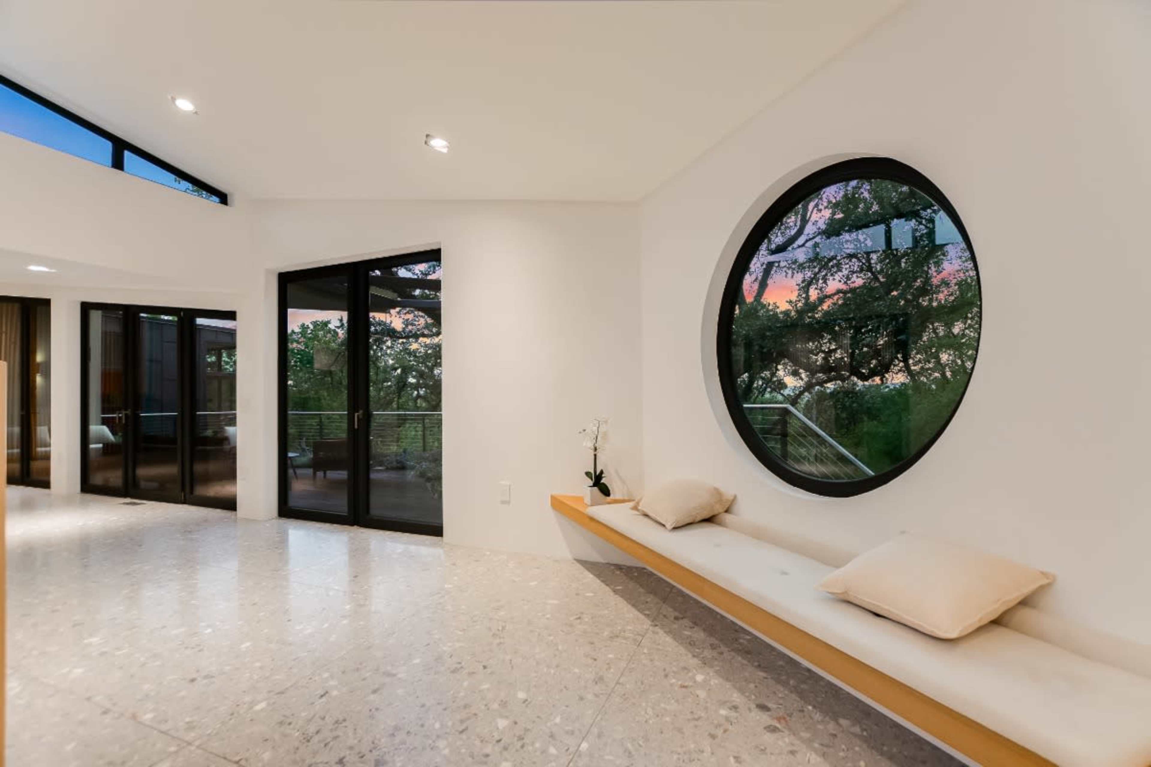 A modern interior features a circular window reflecting trees and a sunset, with a simple bench and two decorative cushions against a white wall.