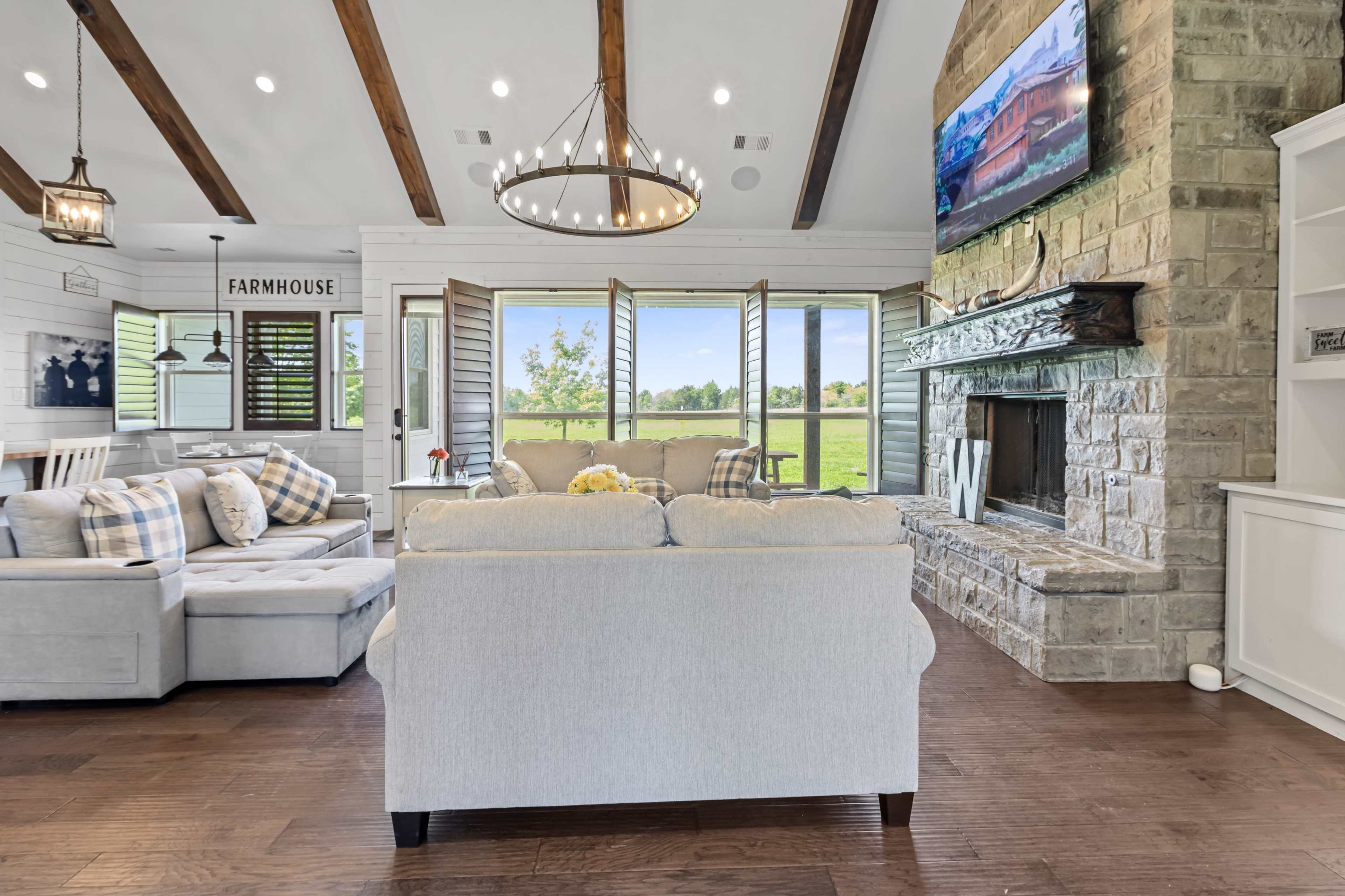 A comfortable living room features a light-colored sofa, a stone fireplace, and large windows overlooking a green outdoor landscape.