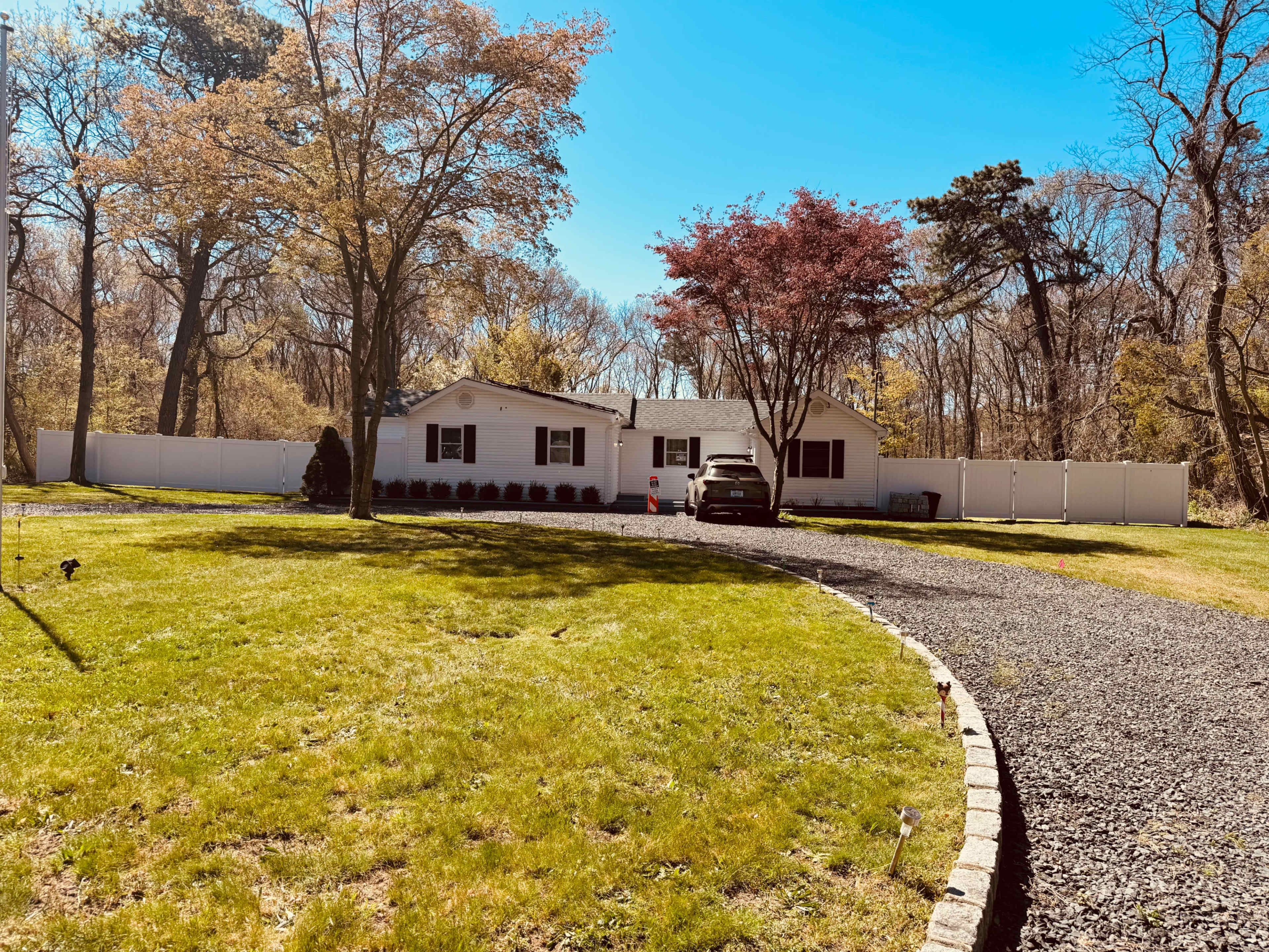 A single-story house with a white exterior is surrounded by a grassy lawn and a gravel driveway, bordered by trees and a white fence.