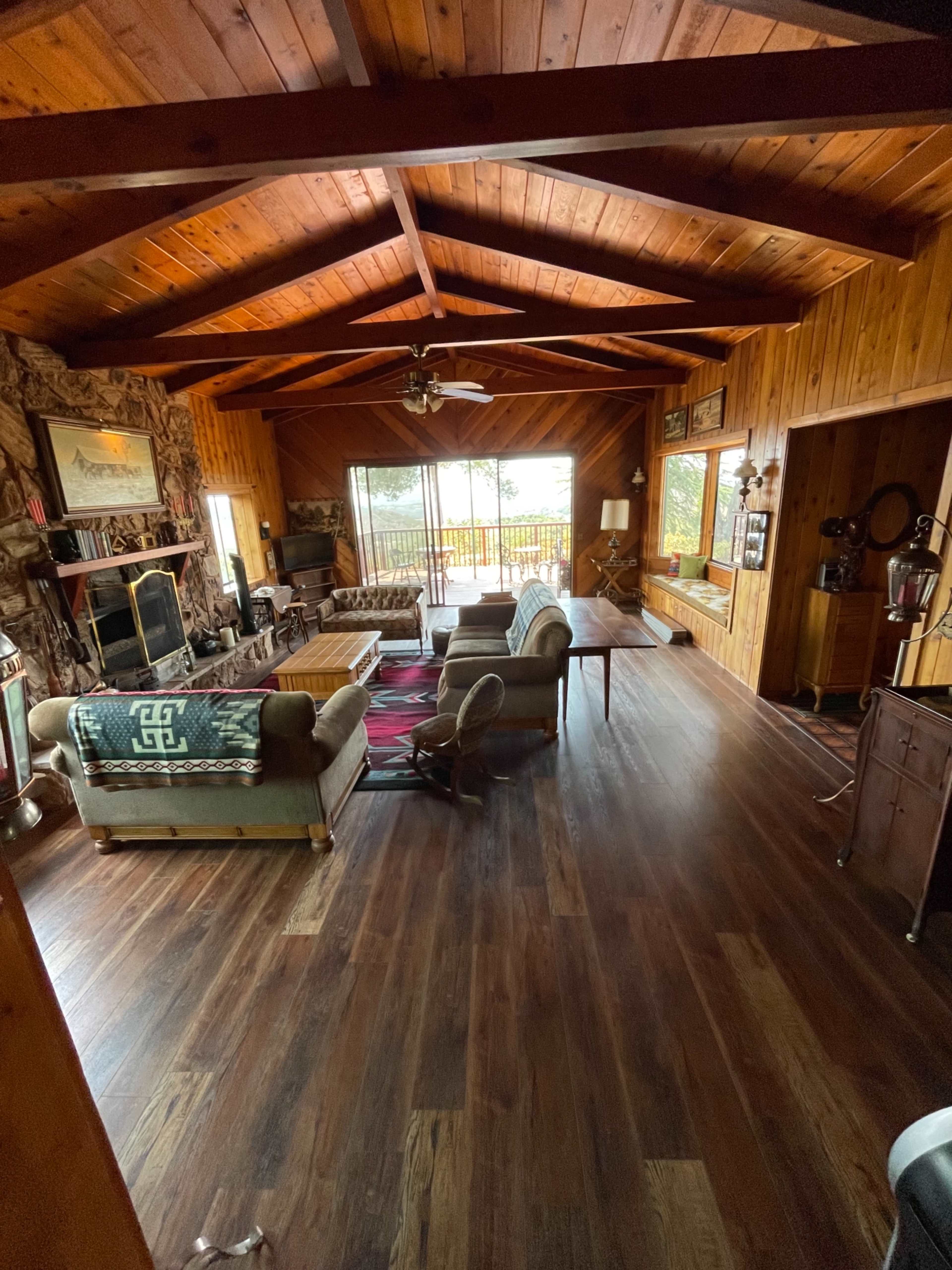 The living room features wooden walls and a high-beamed ceiling, with a central seating area arranged around a large window overlooking a deck.