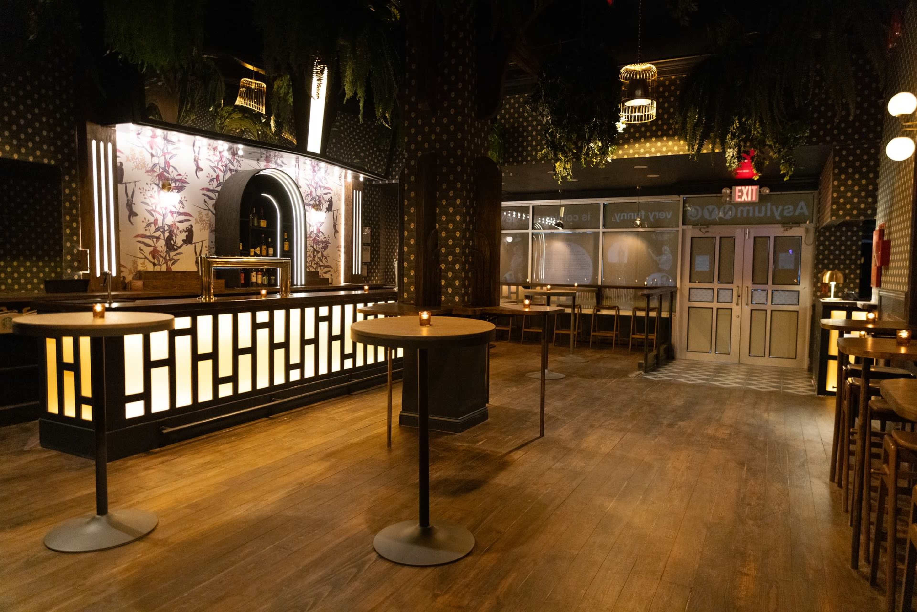 A dimly lit restaurant interior featuring a bar with a decorative backdrop and wooden tables arranged throughout the space.