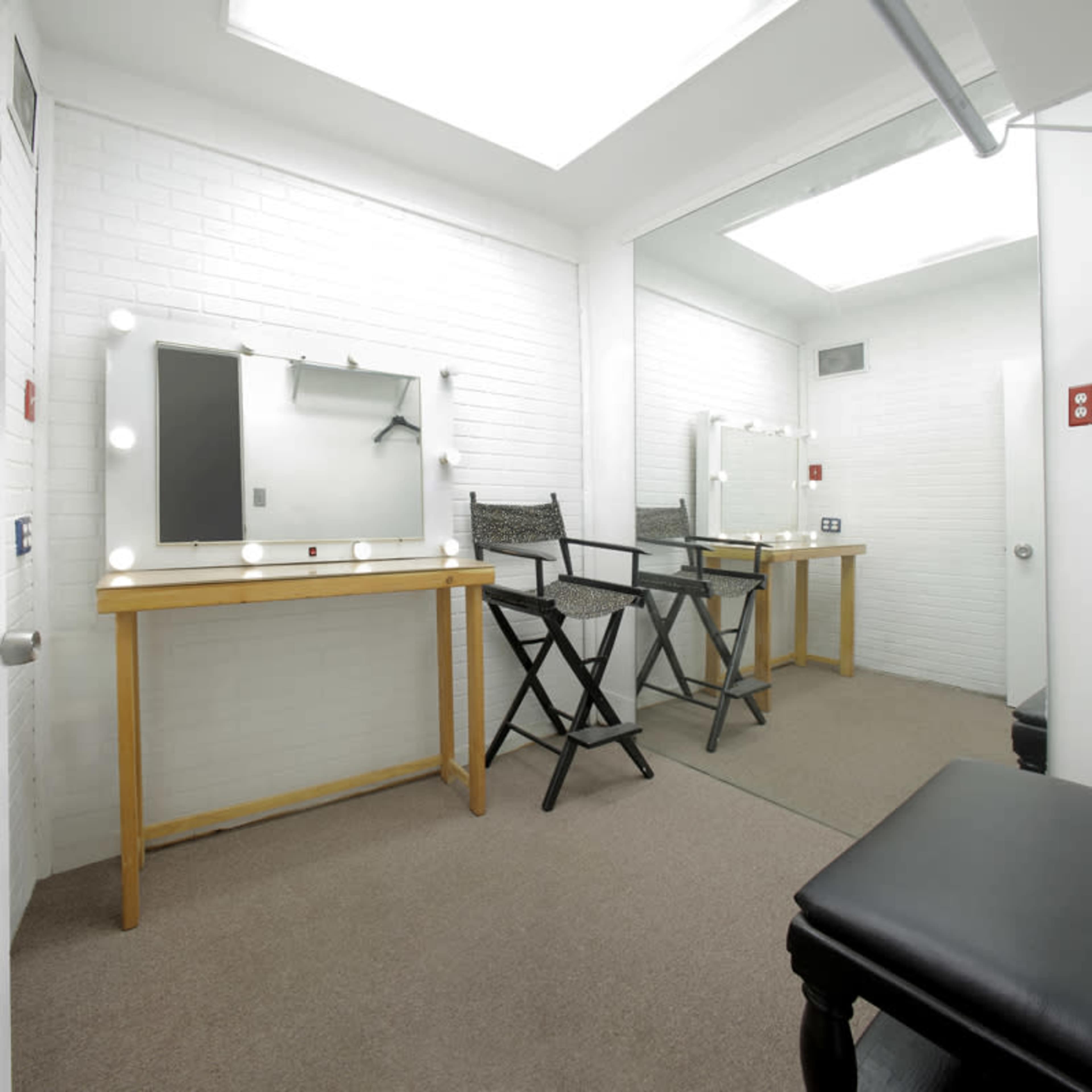 The image shows a well-lit dressing room with a large mirror surrounded by lights, two wooden stools, and a black bench against a white-brick wall.