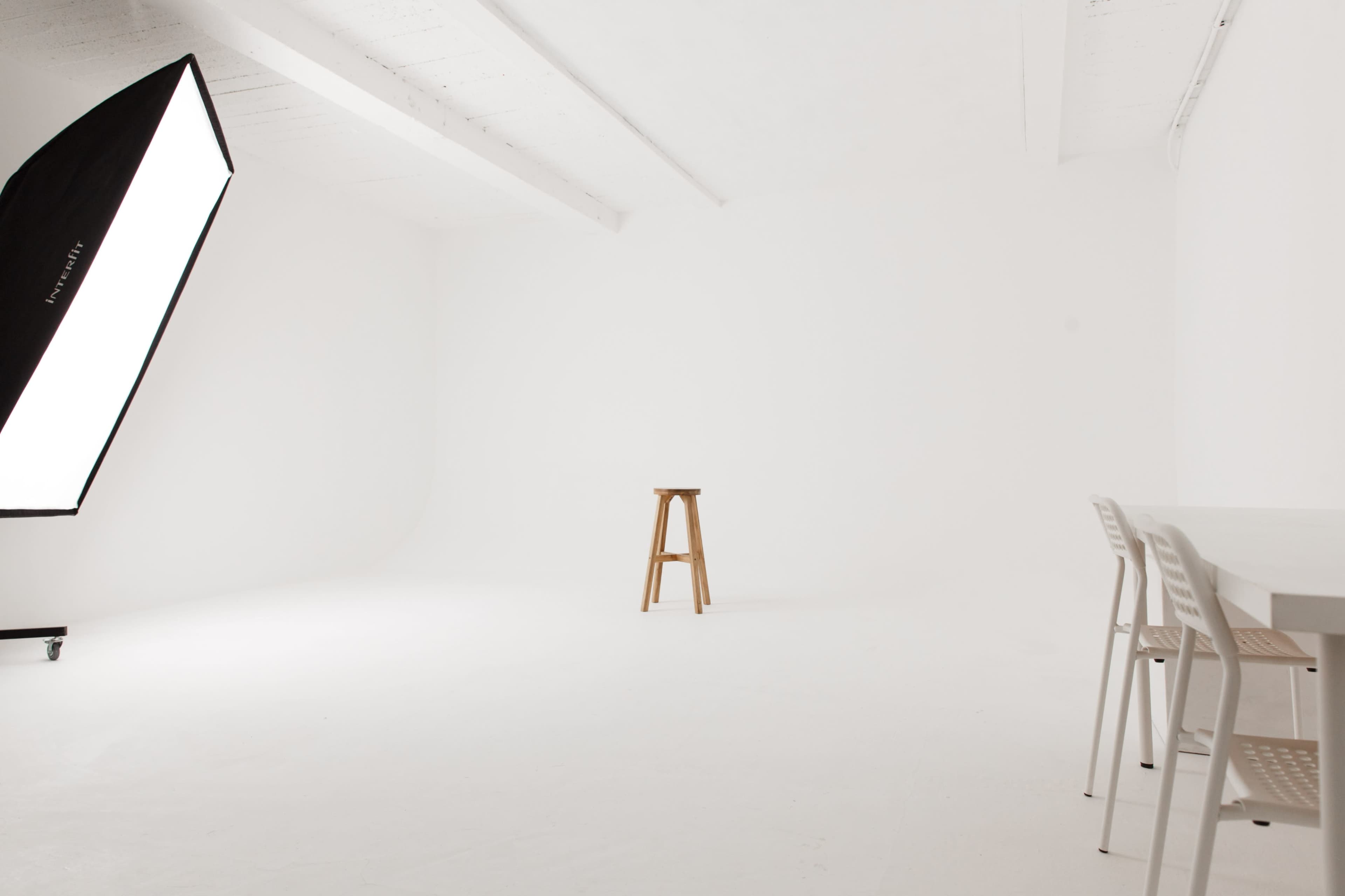 A wooden stool is positioned in a minimalistic white room with a large softbox light and a set of white chairs nearby.