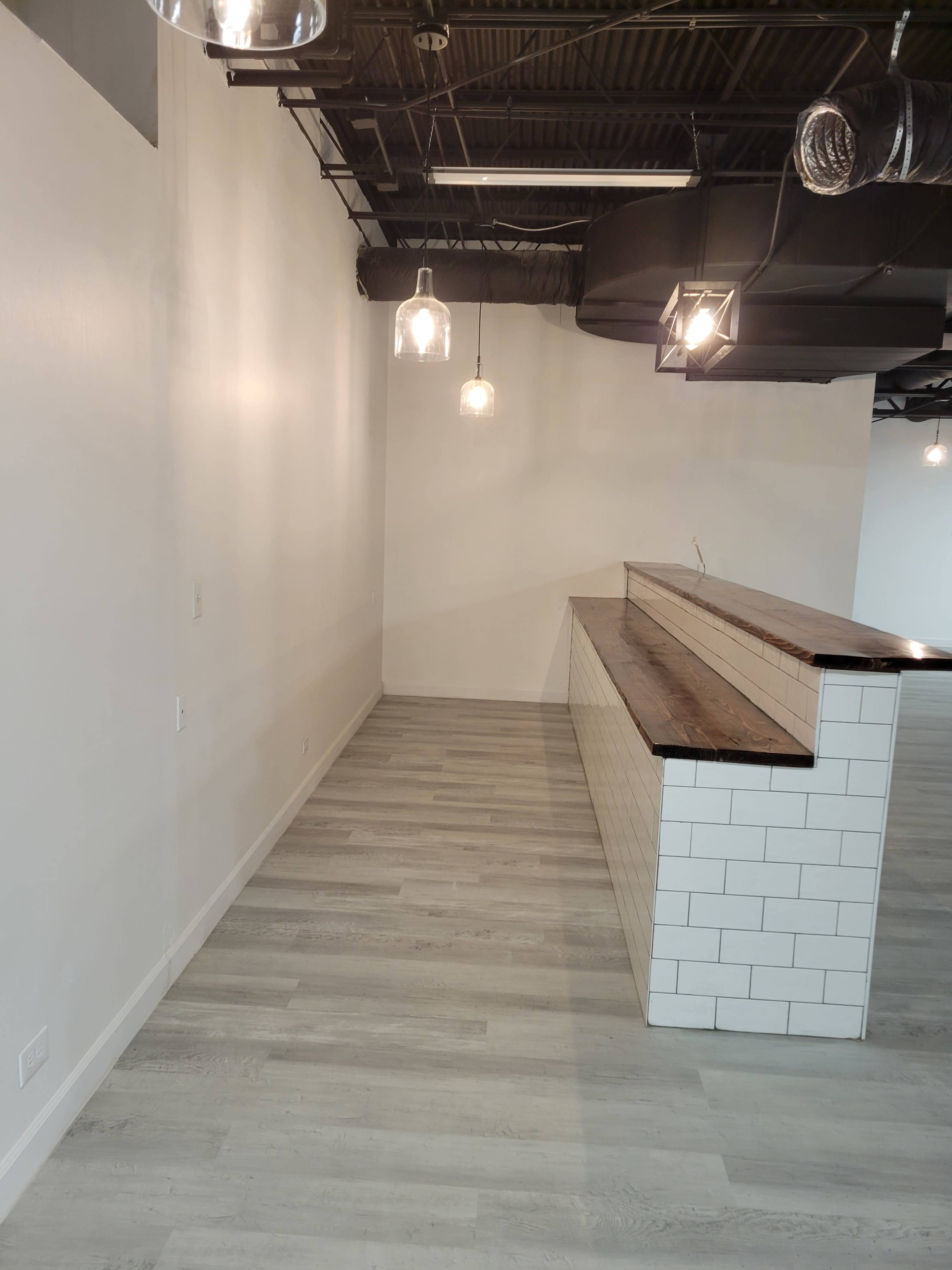 The image shows a modern, vacant interior space featuring a wooden counter with white tiled accents and pendant lighting.
