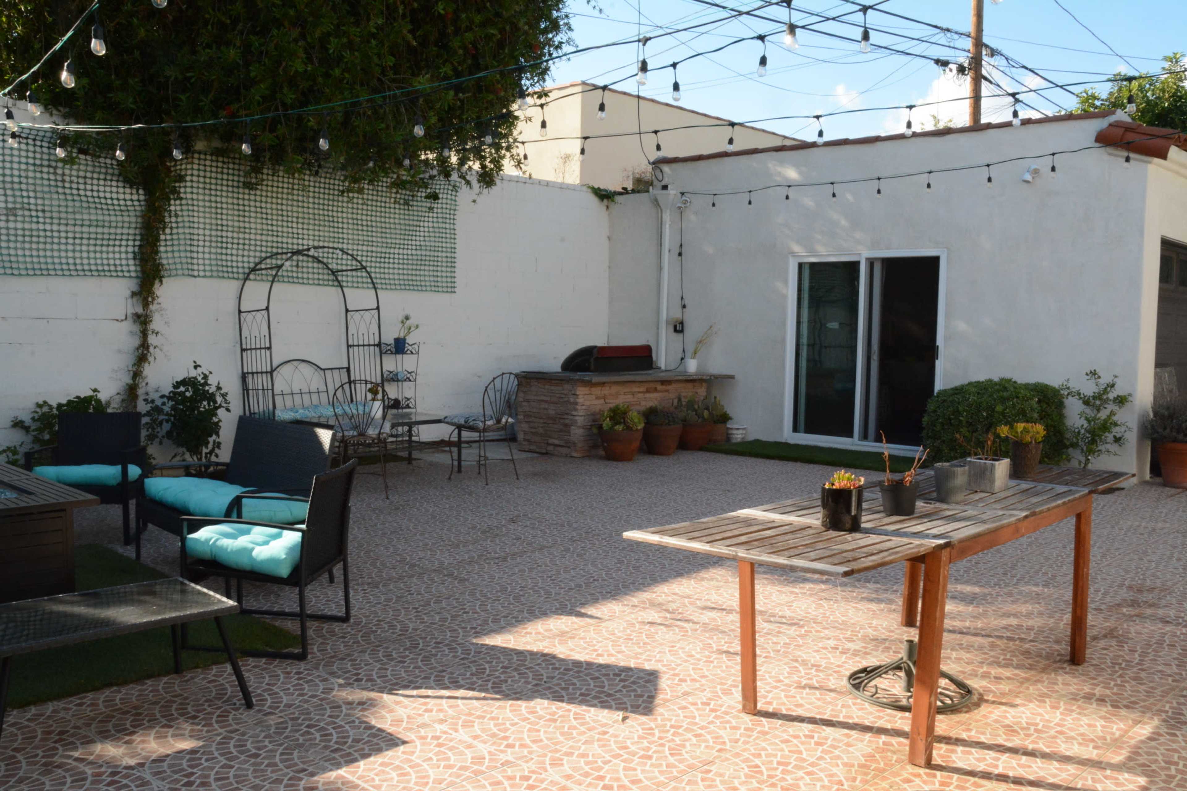 The outdoor space features a paved patio with seating arrangements, a wooden table with potted plants, and string lights overhead.