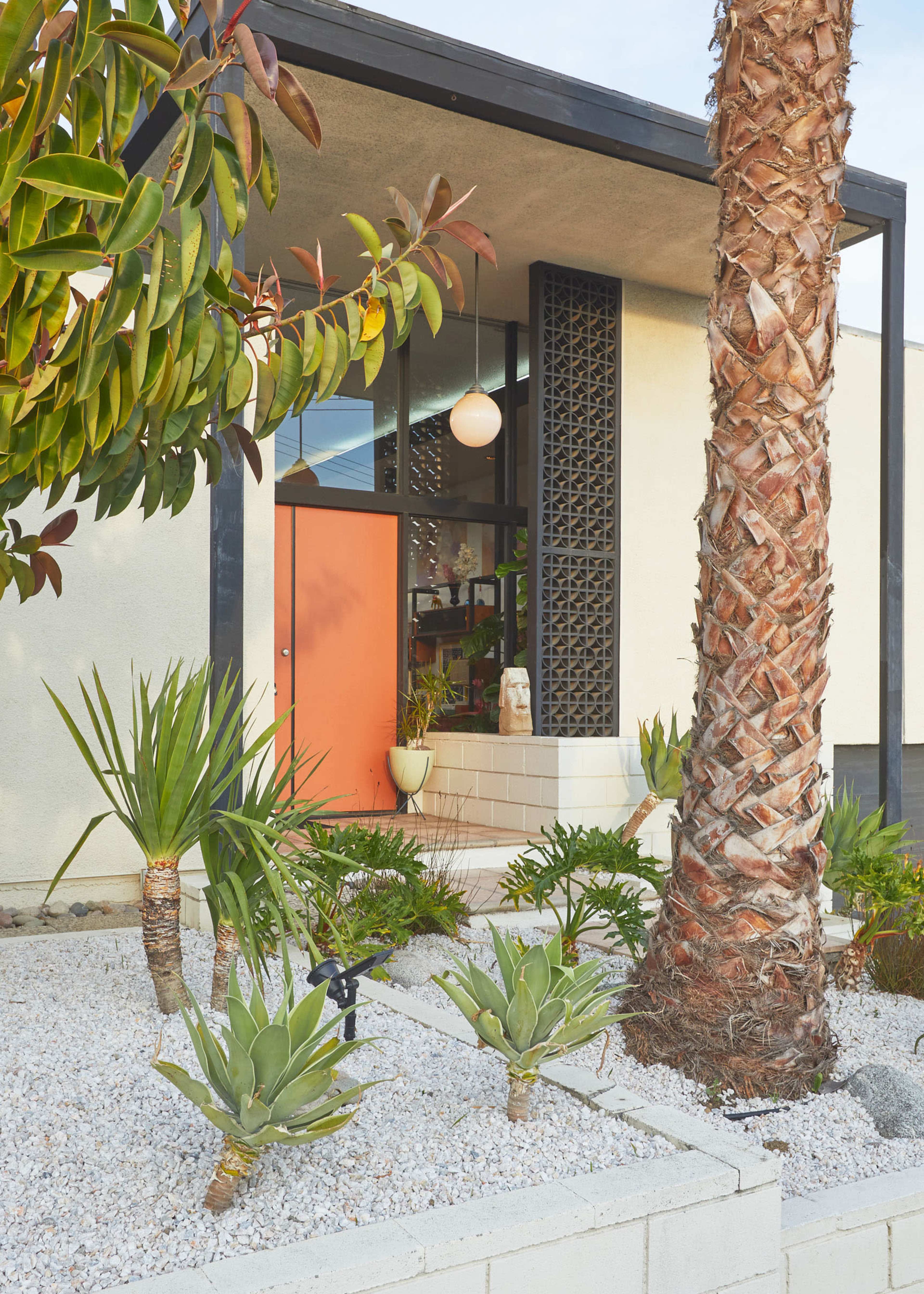 A modern home features an orange door, surrounded by landscaped gravel and various plants, including agaves and a palm tree.