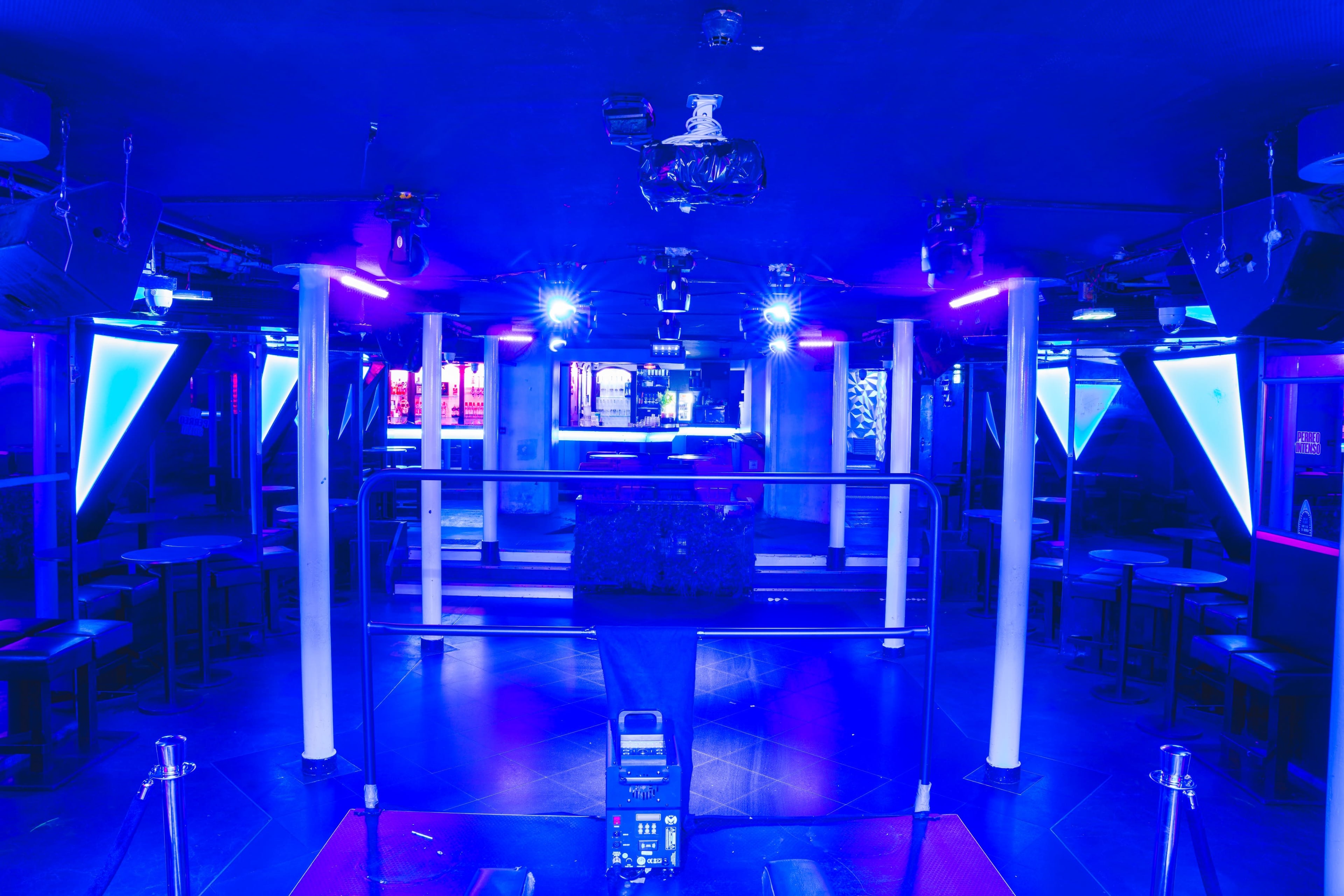 A dimly lit nightclub features blue and purple lighting, empty seating, and a central stage area.