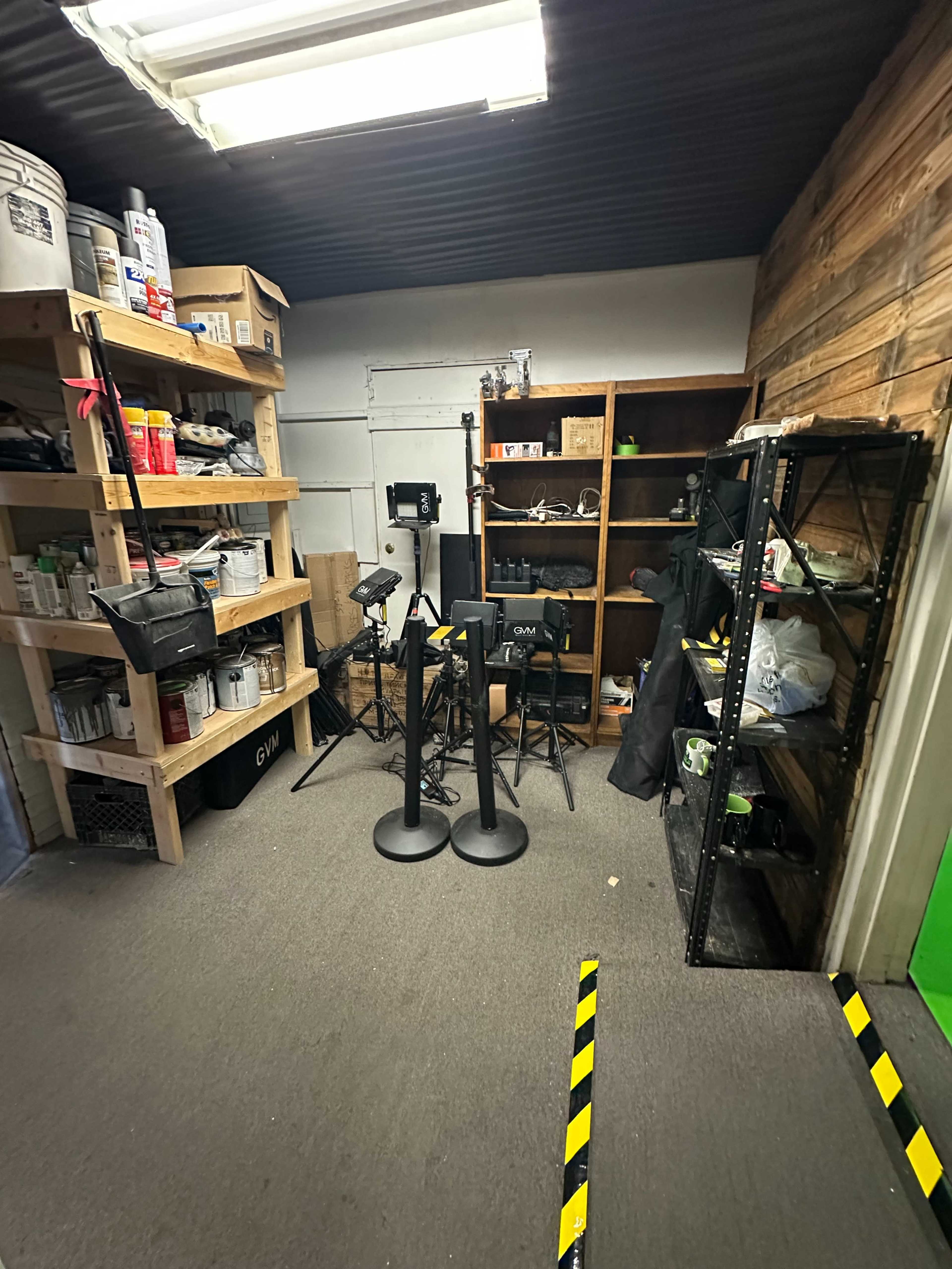 The image shows a small storage room with shelving units filled with various tools and supplies, along with a couple of stands and some chairs positioned nearby.