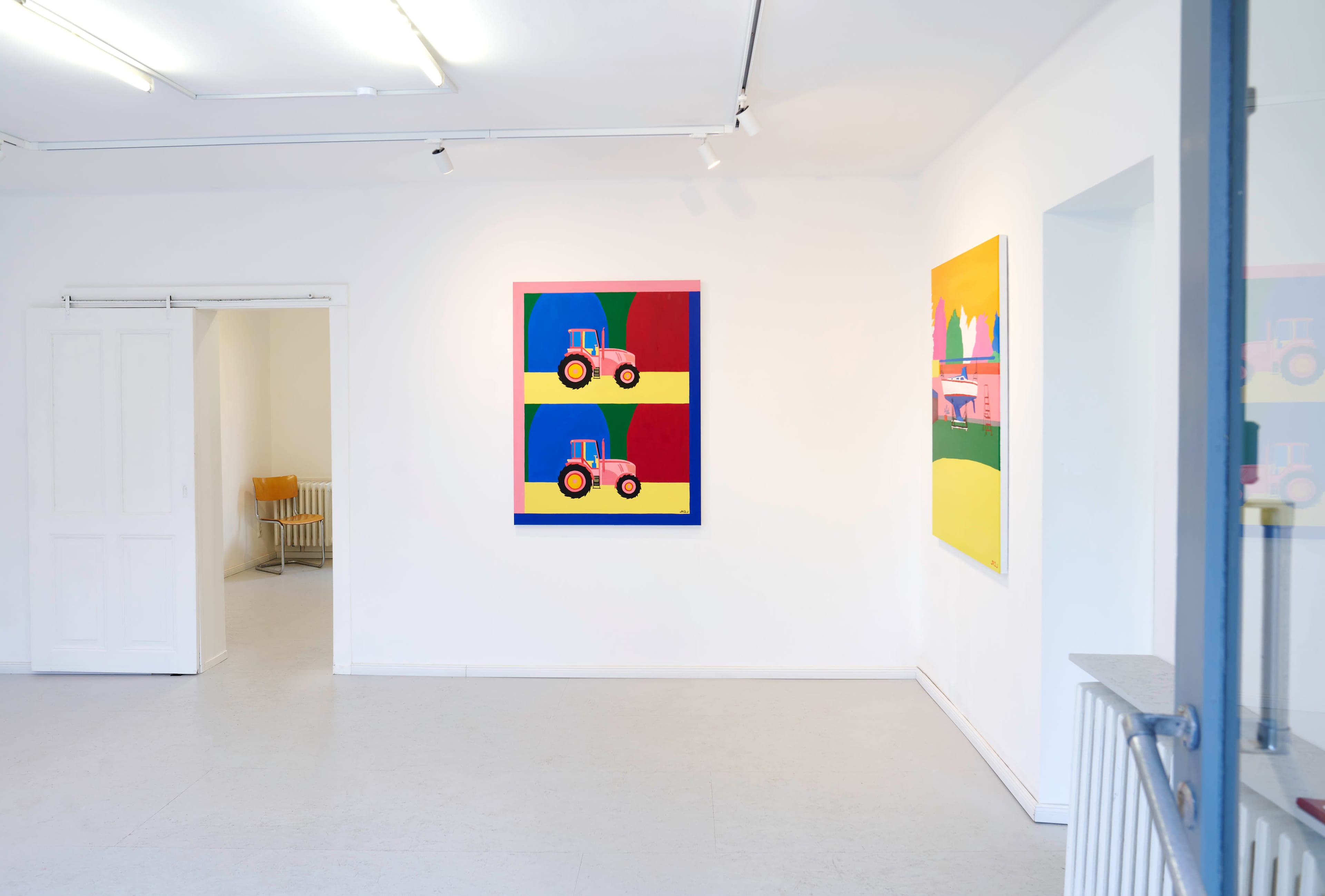 A bright art gallery features two colorful abstract paintings on white walls, with a visible doorway leading to a separate room and a chair in the background.