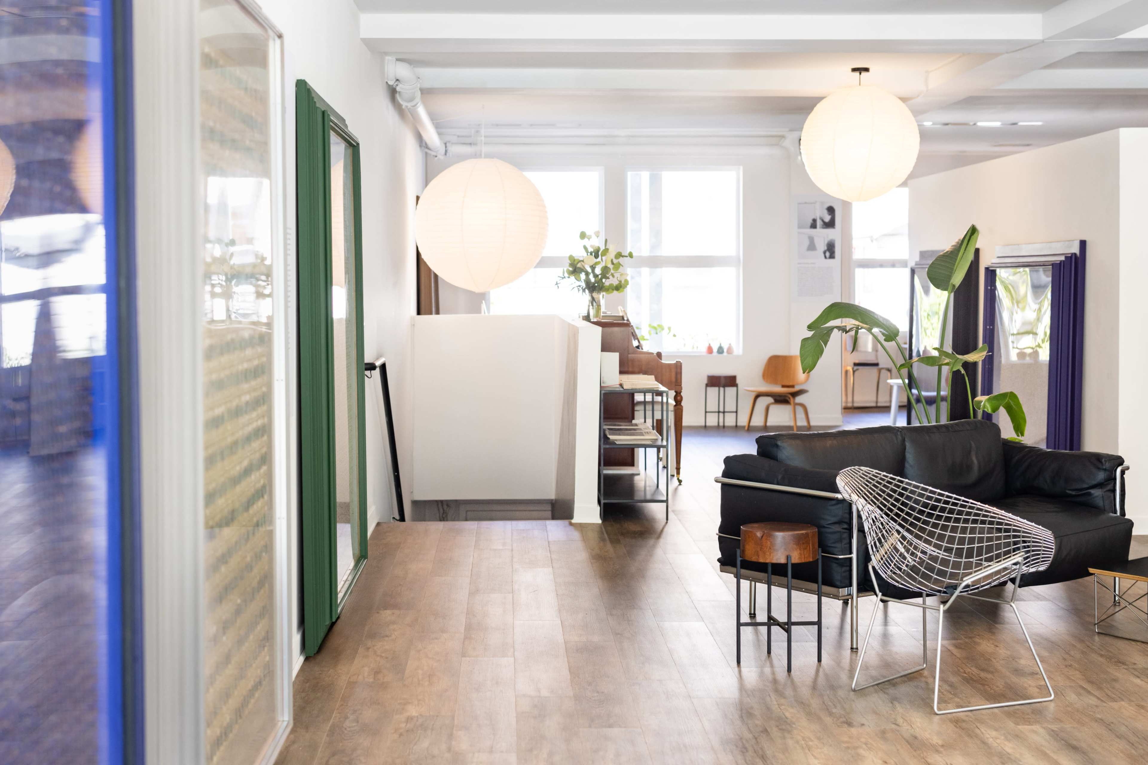 The image shows a modern interior space with wooden flooring, a black sofa, a wire chair, and large paper lanterns hanging from the ceiling.