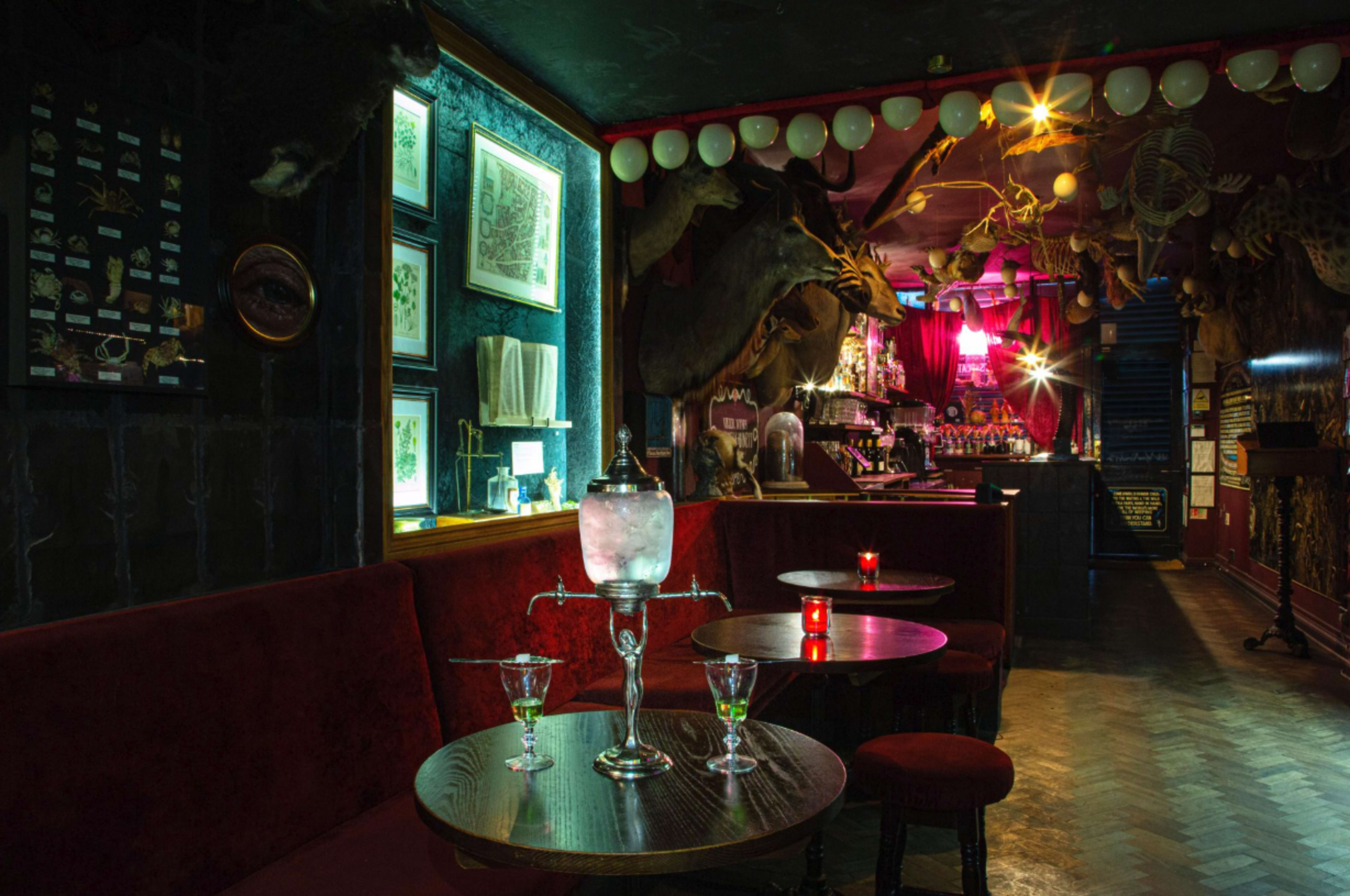 The image depicts a dimly lit bar with plush red seating, adorned with animal mounts and eclectic decorations, featuring tables with drinks and a bar area in the background.