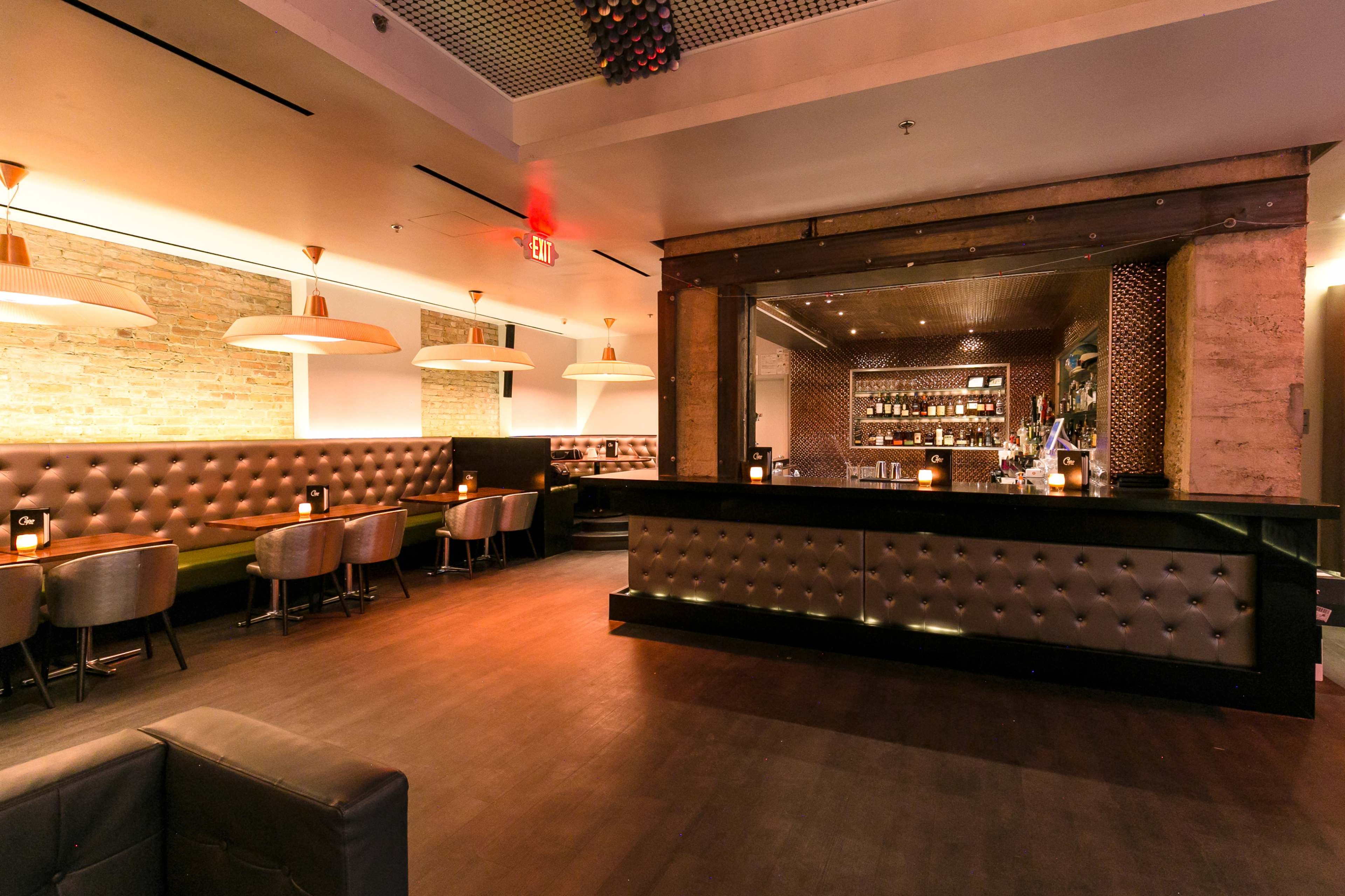 An empty bar area with tufted seating, pendant lights, and a sleek counter against a backdrop of exposed brick.