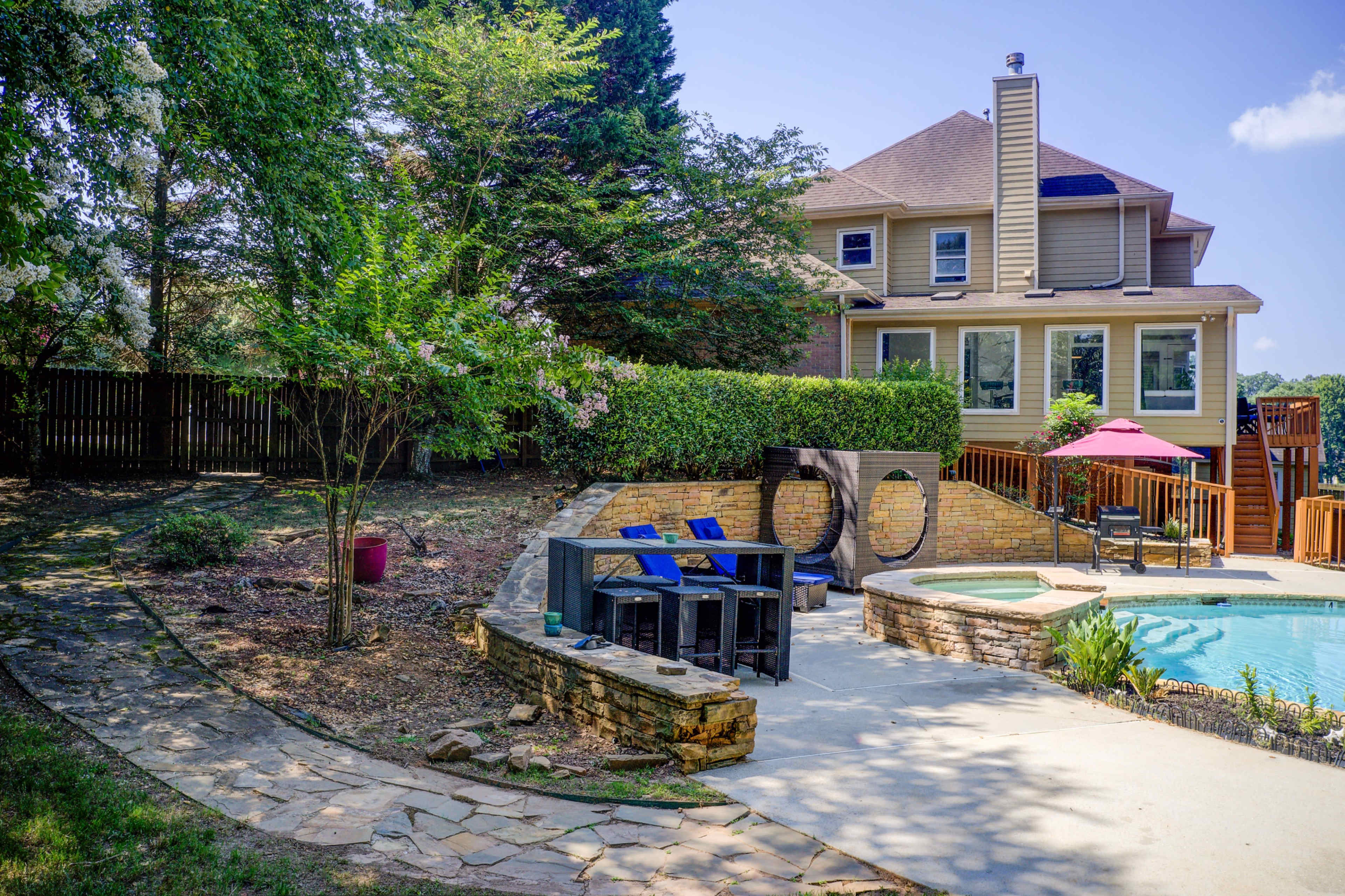 A backyard features a swimming pool, stone patio, and a landscaped area with trees, beside a two-story house.