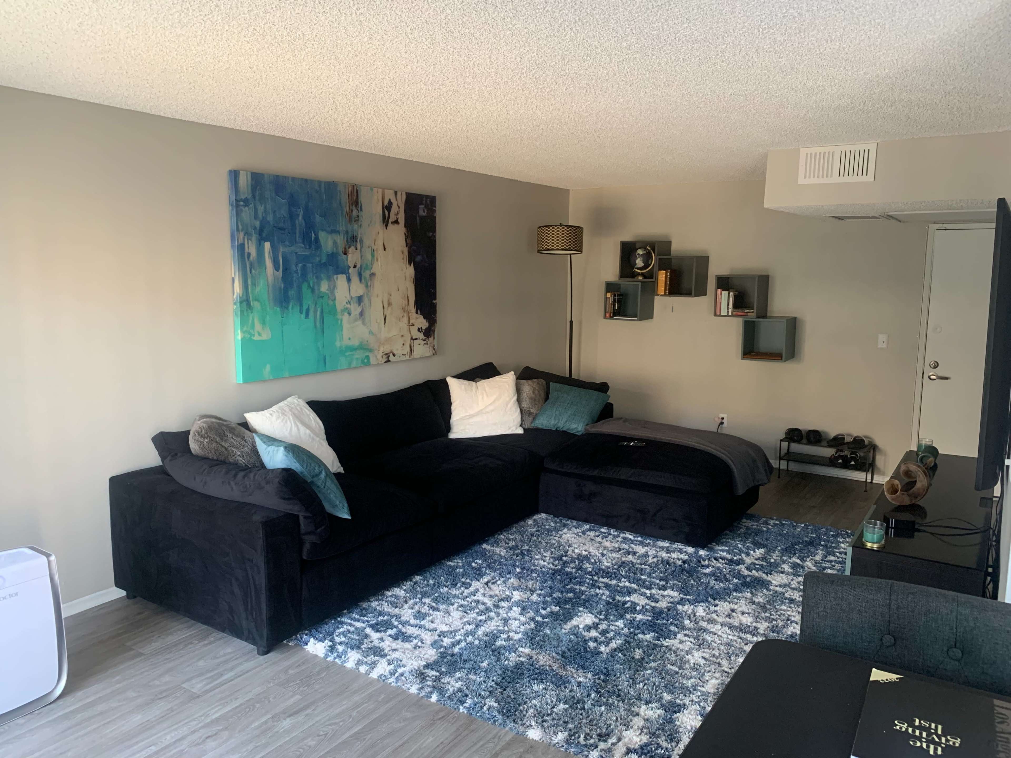 A modern living room with a black sectional sofa, a blue abstract painting on the wall, and a bookshelf with decorative items.
