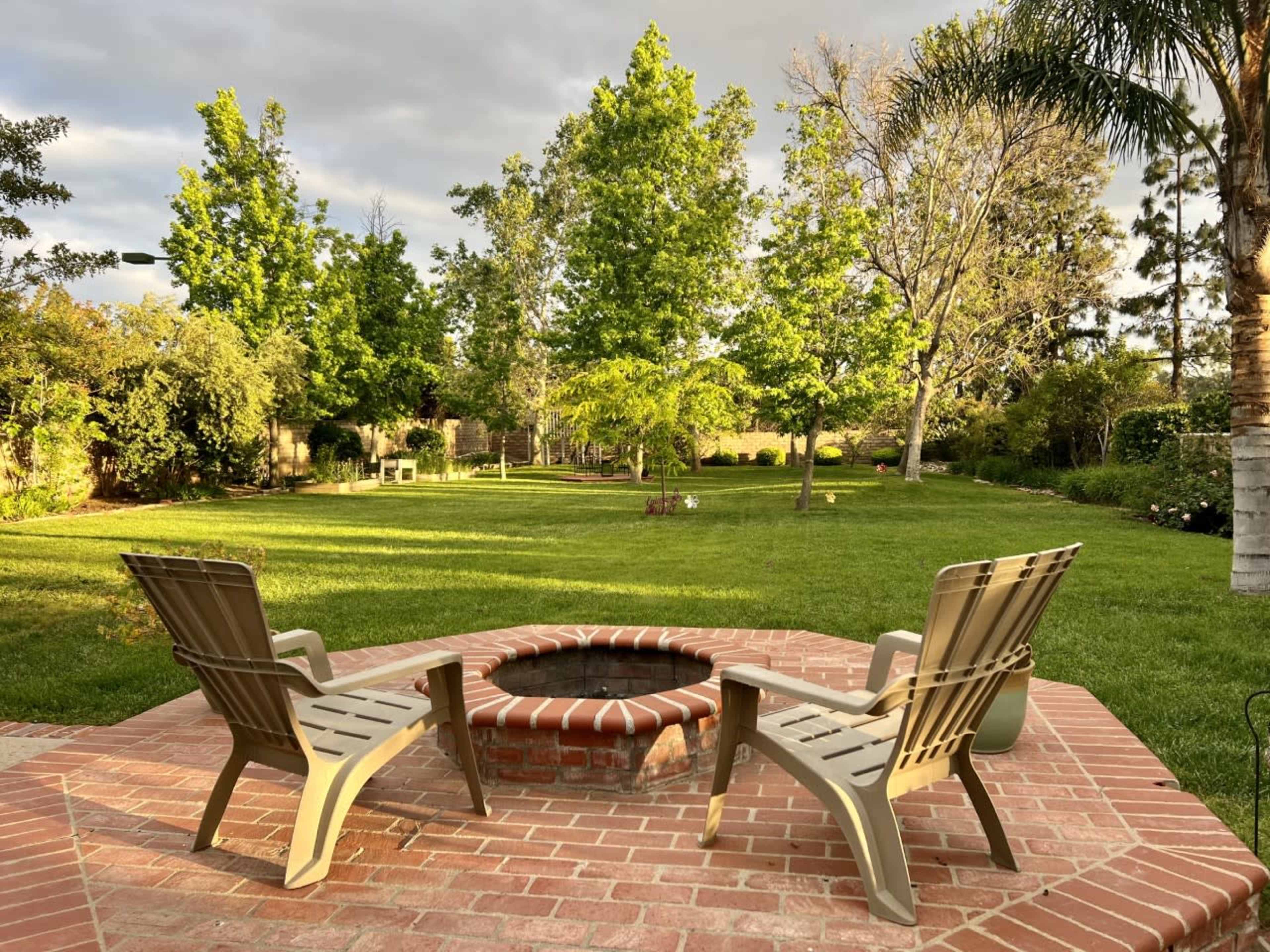Two chairs positioned around a circular brick fire pit in a grassy backyard with trees and shrubs.