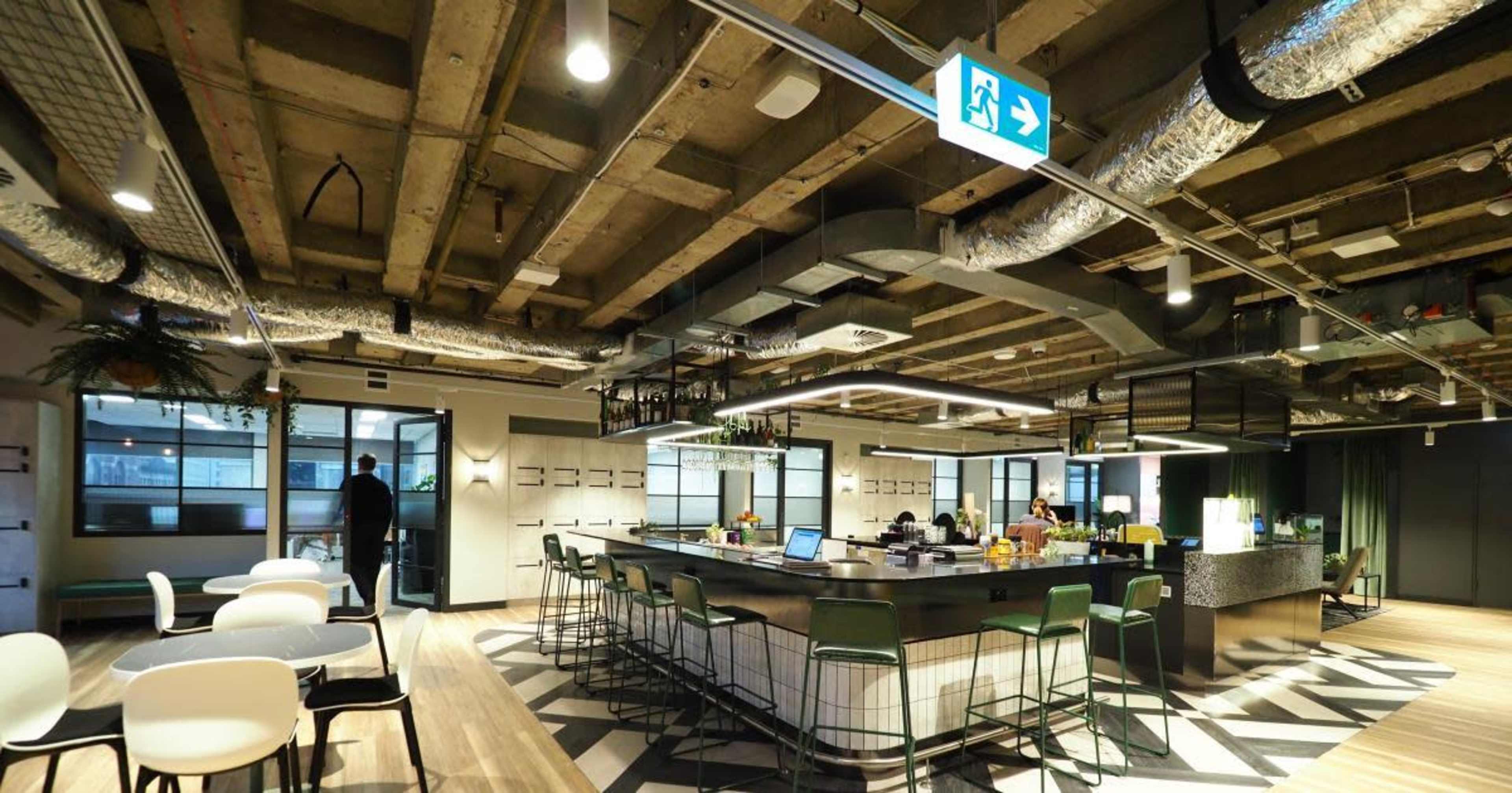 A modern office space featuring a bar area with high stools, tables, and people working on laptops under exposed ceiling beams.