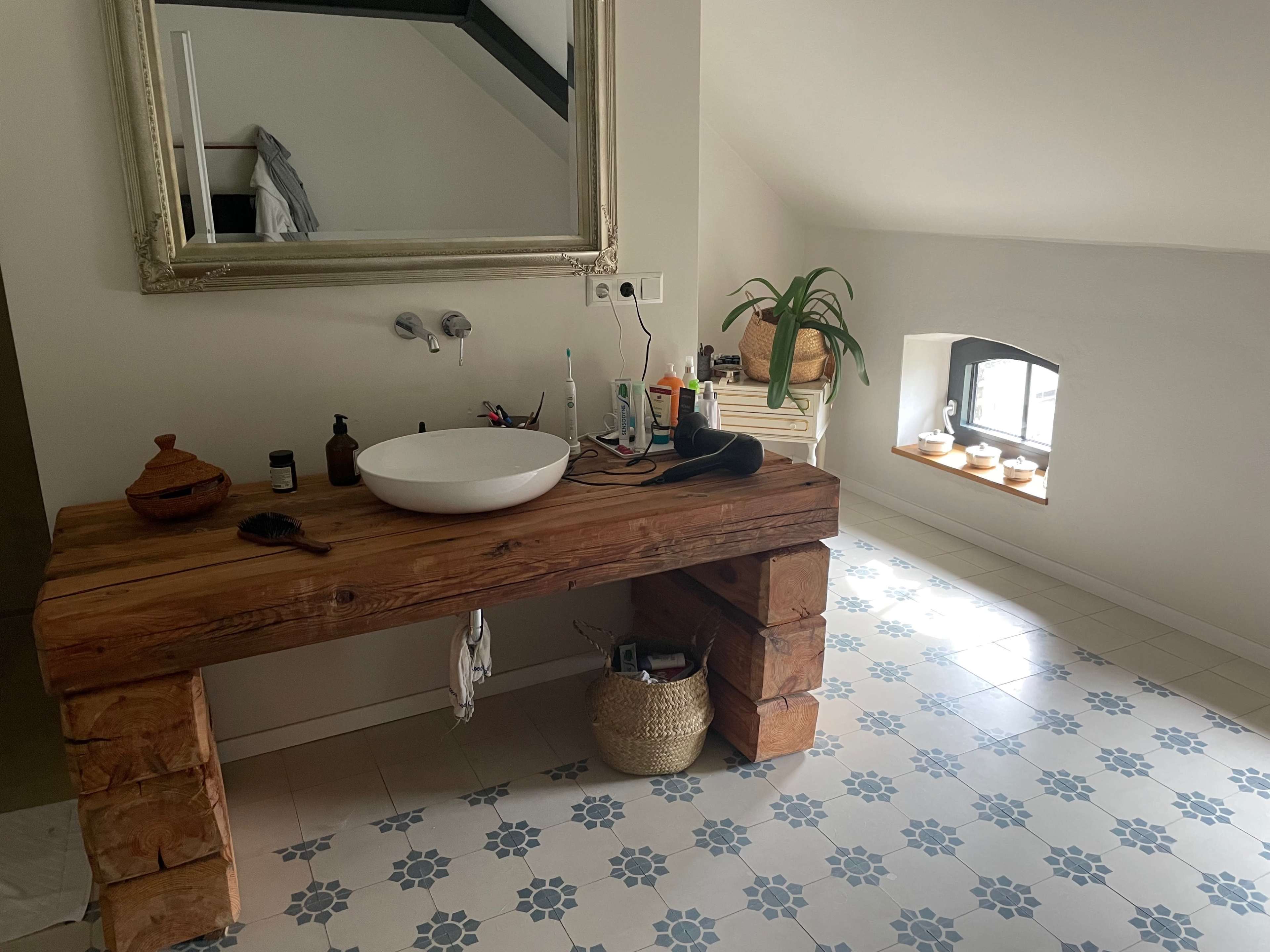 The image shows a bathroom featuring a wooden sink vanity with a round basin, a decorative mirror above it, and a window with potted plants beside it.