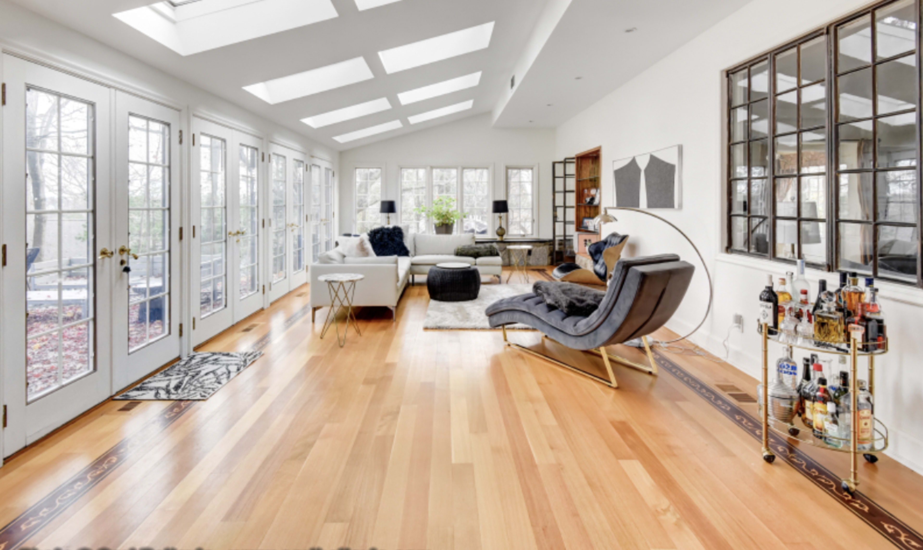 A brightly lit sunroom with large glass doors, hardwood flooring, and modern furniture, including a stylish chaise lounge and a bar cart.