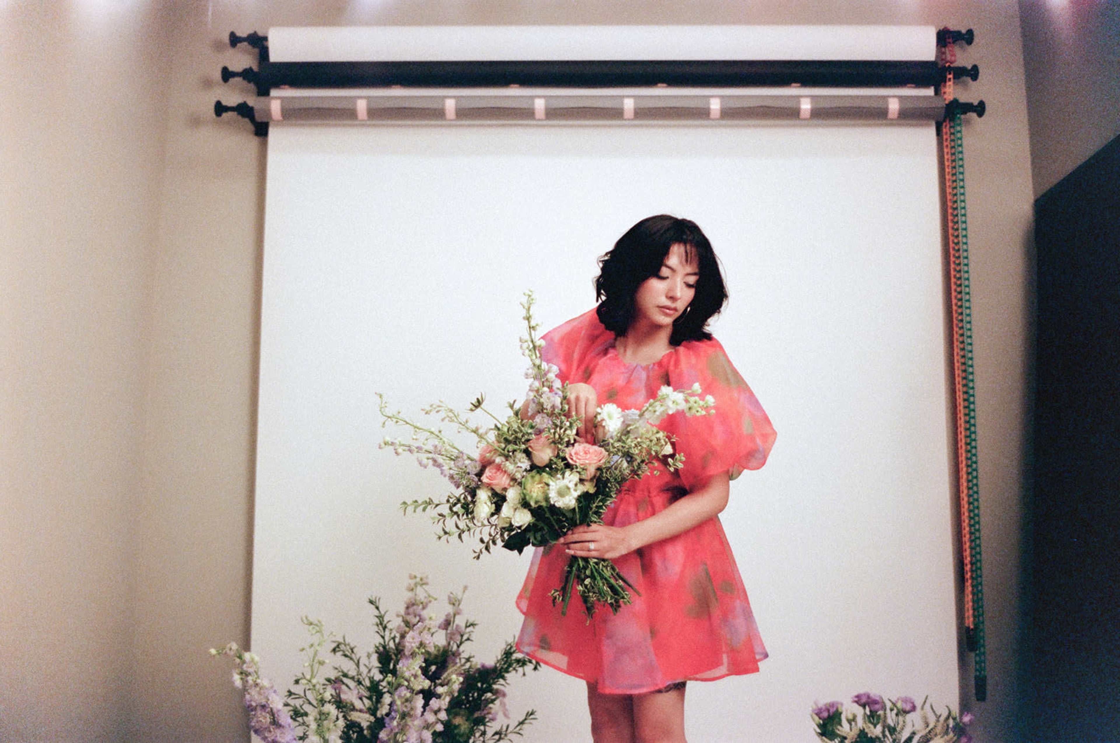 A woman in a pink dress holds a large bouquet of flowers in front of a plain backdrop.