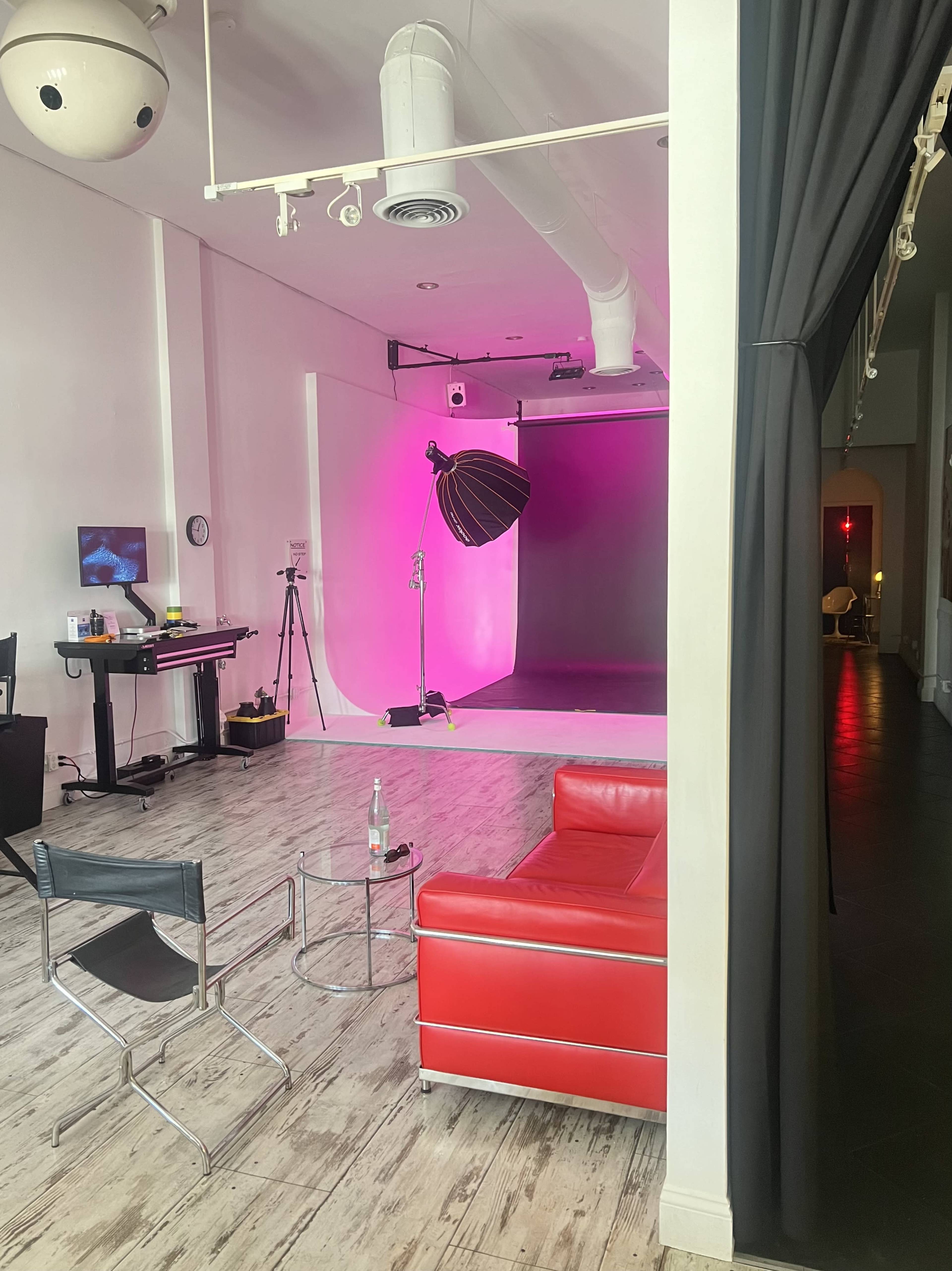 The image shows a studio space featuring a red couch, a lighting setup with a softbox, and a pink backdrop along with various equipment.