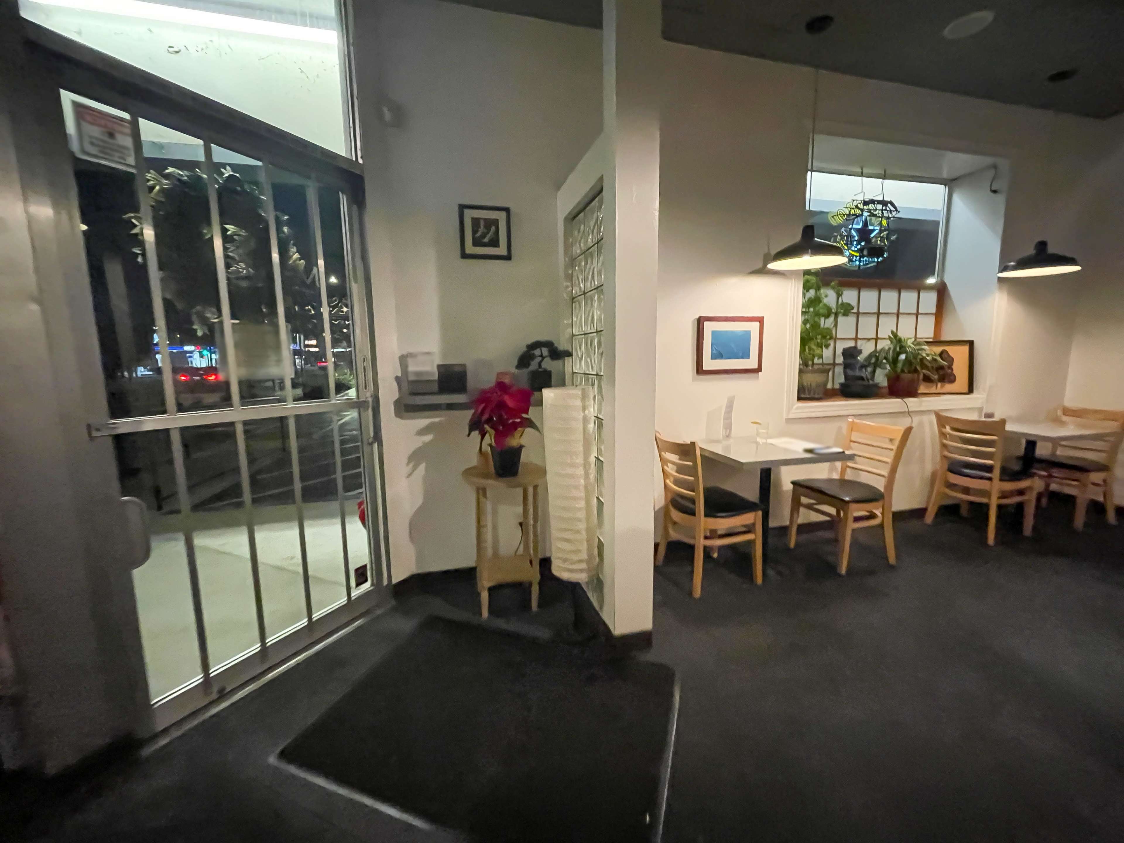 The image shows a dimly lit restaurant interior with a decorative plant on a small table, wooden chairs, and artwork on the walls.