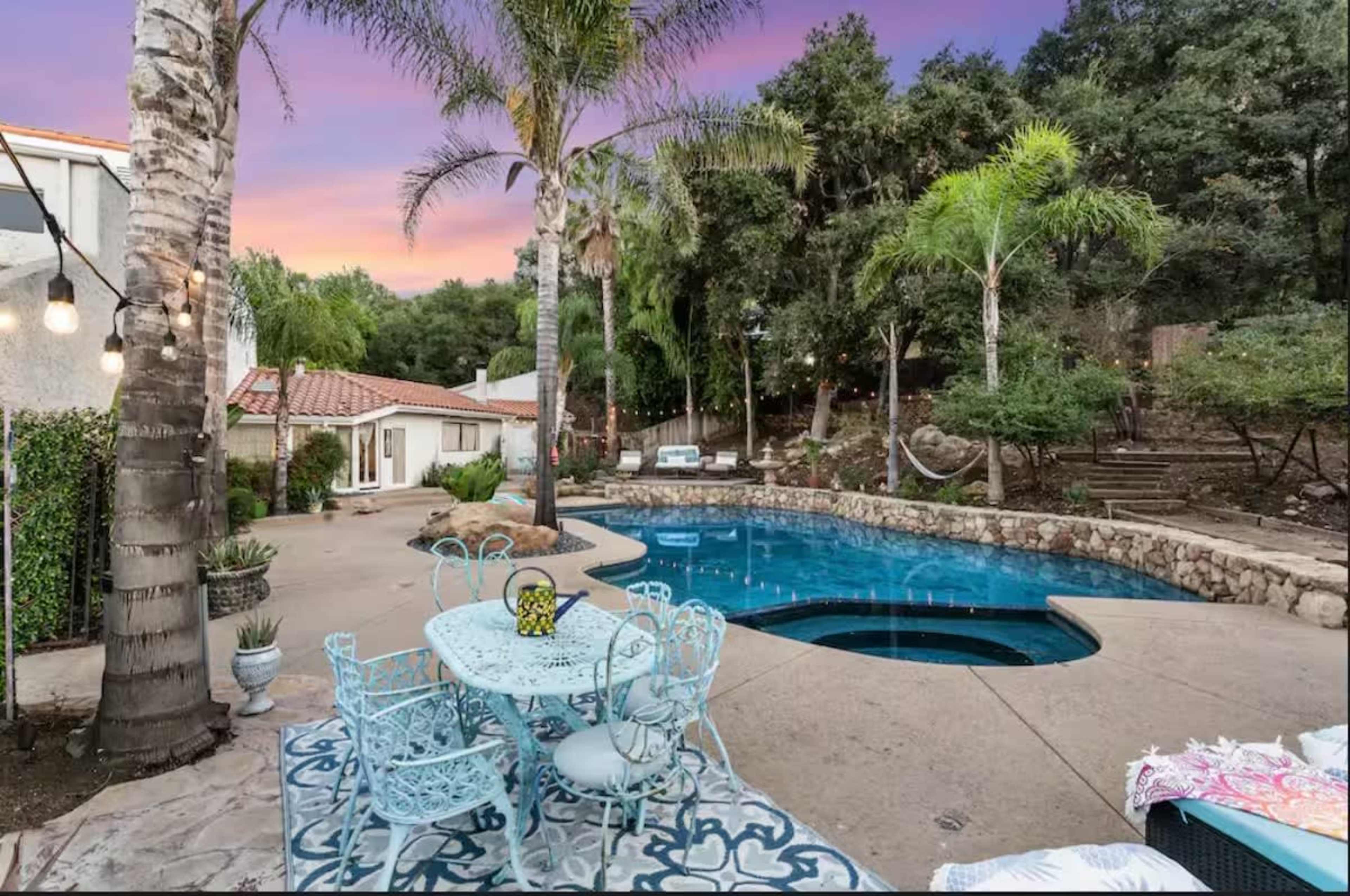 The image shows a backyard with a swimming pool surrounded by palm trees, a stone wall, and a dining area with a light blue table and chairs.