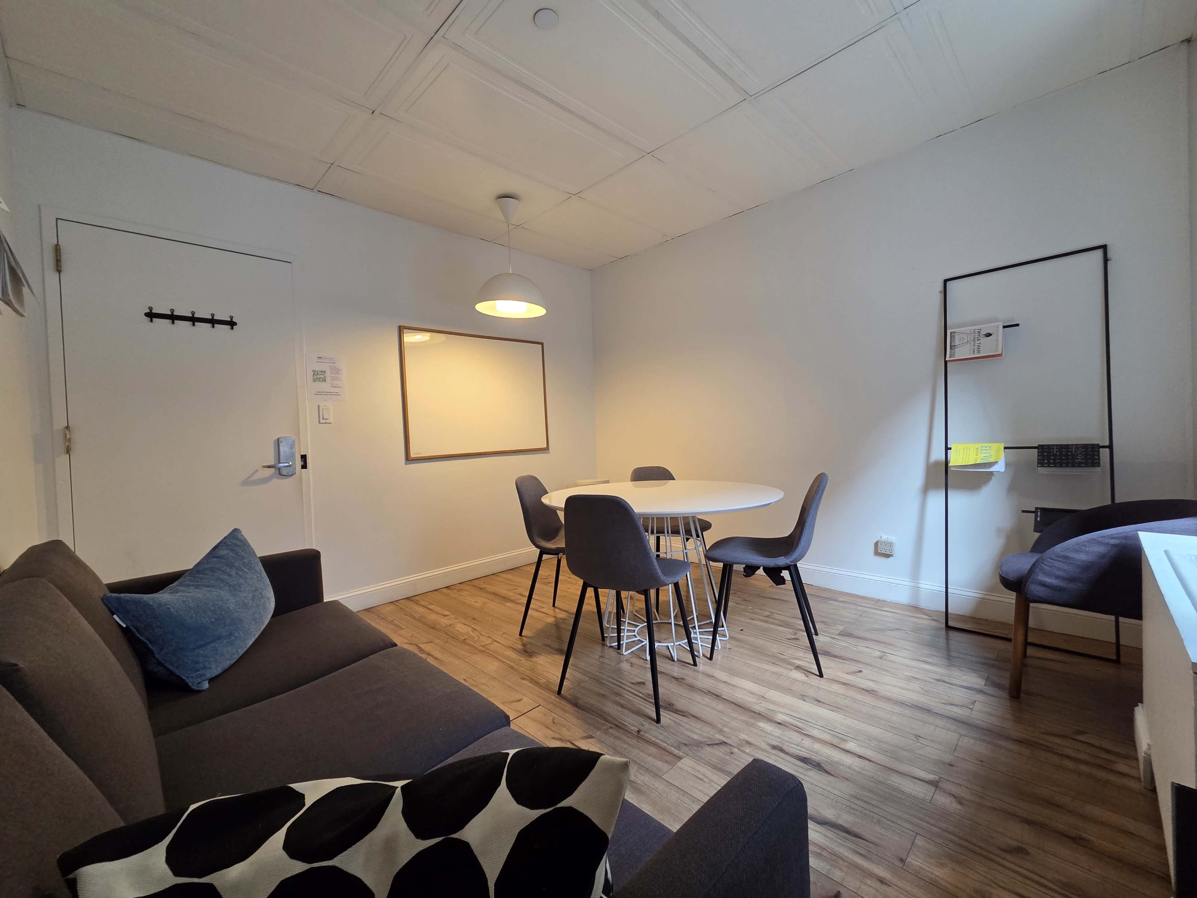 The image shows a small, modern meeting room with a round white table surrounded by six chairs, a gray sofa with a blue cushion, and a whiteboard on the wall.