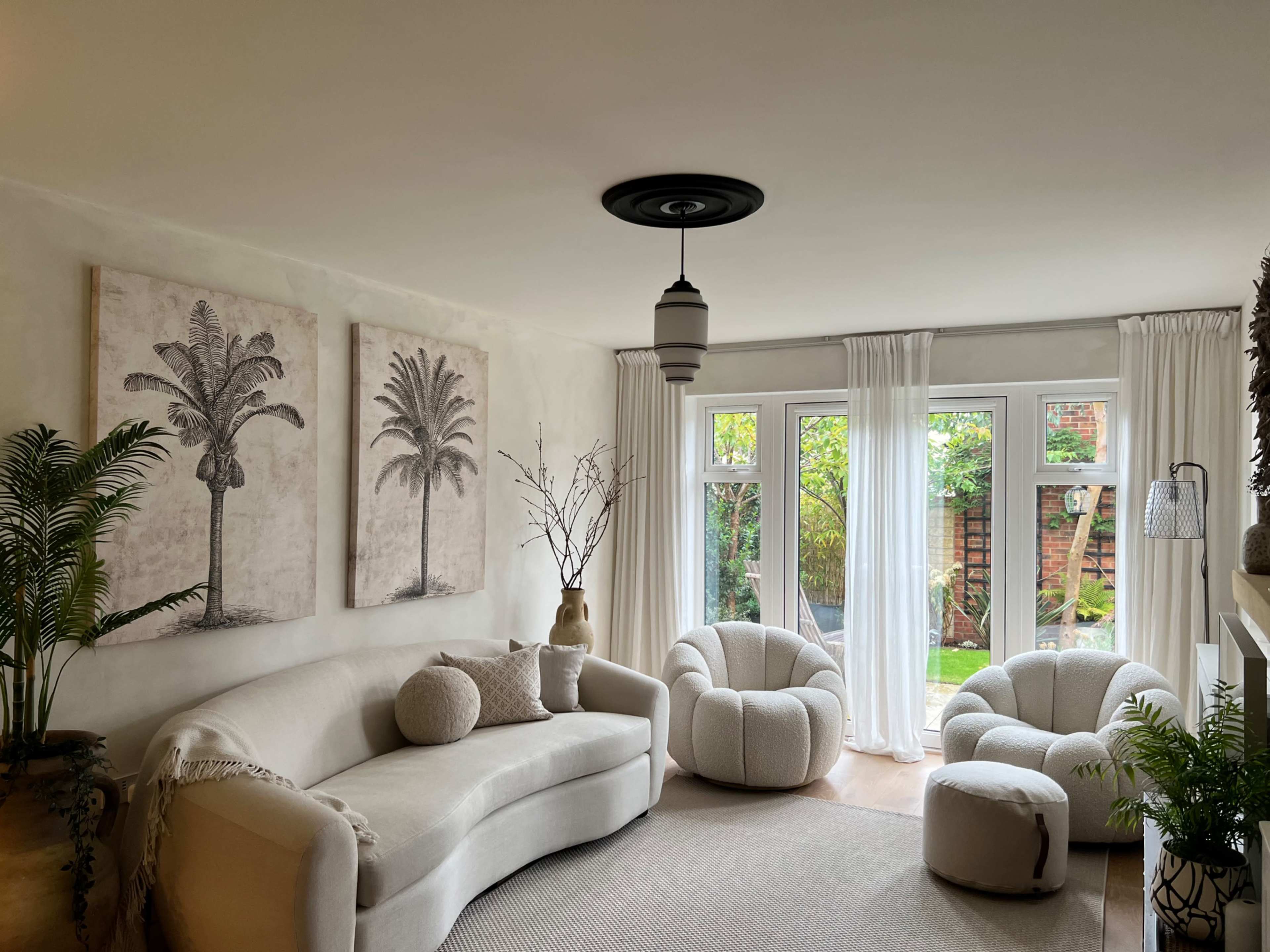 A modern living room featuring a curved sofa, two round armchairs, large windows with sheer curtains, and palm tree artwork on the walls.