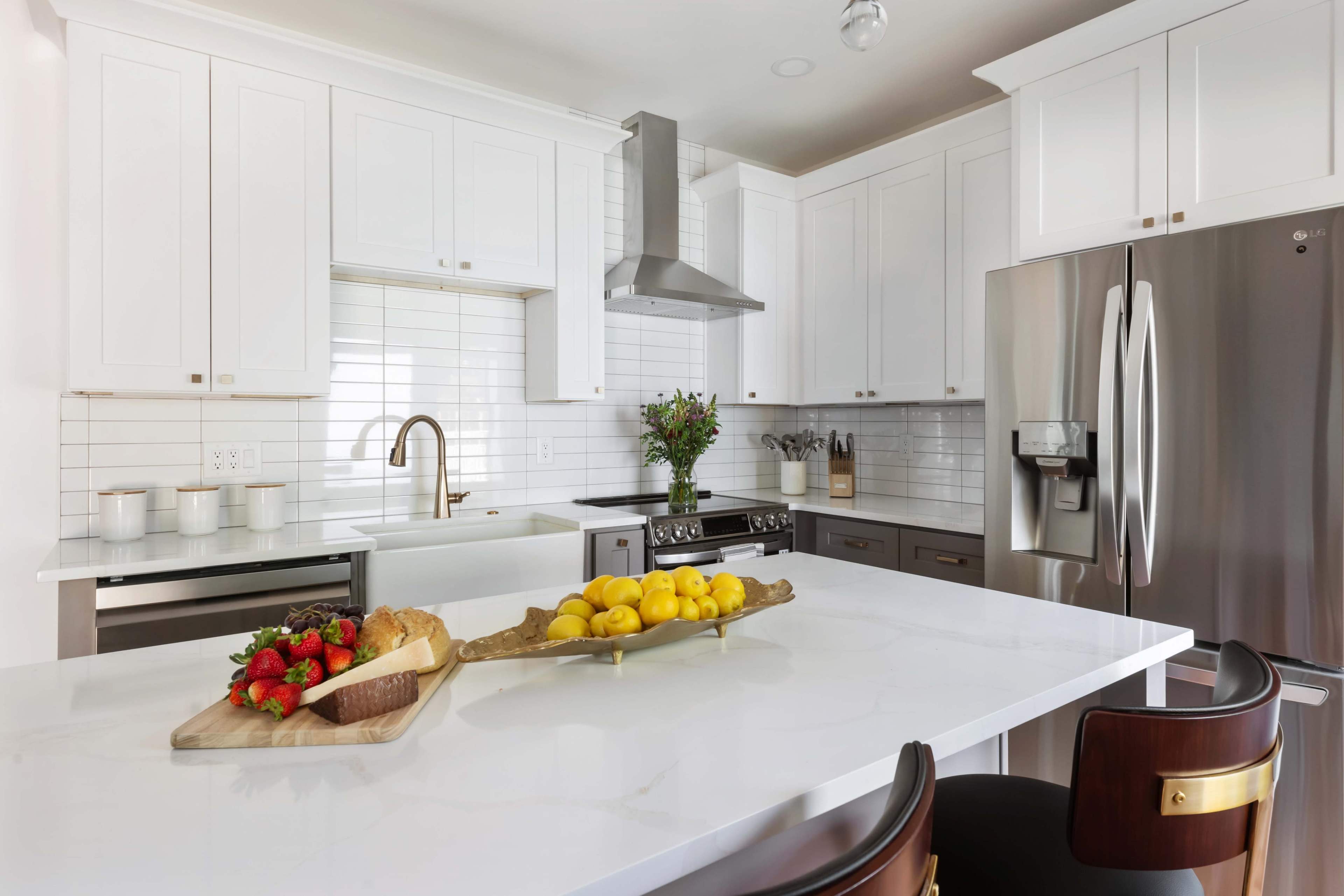 A modern kitchen features white cabinetry, stainless steel appliances, and a central island with a wooden platter holding fruits.
